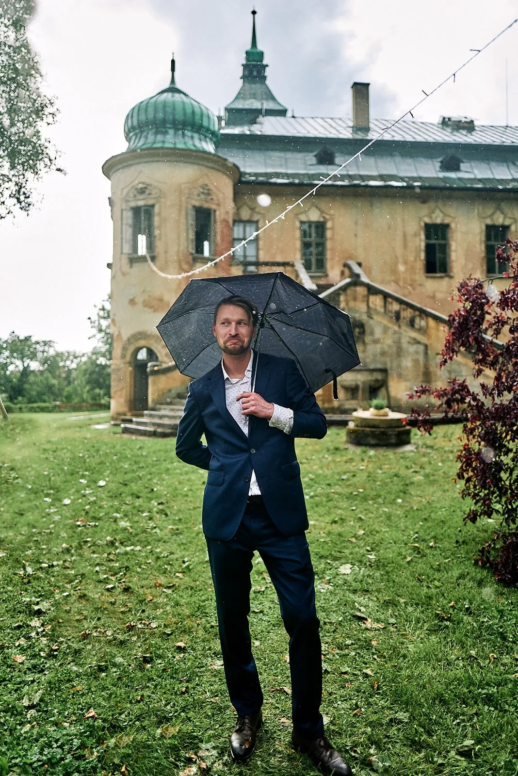 A man in a suit holding a black umbrella, standing on grass in front of an old castle-like building on a rainy day.