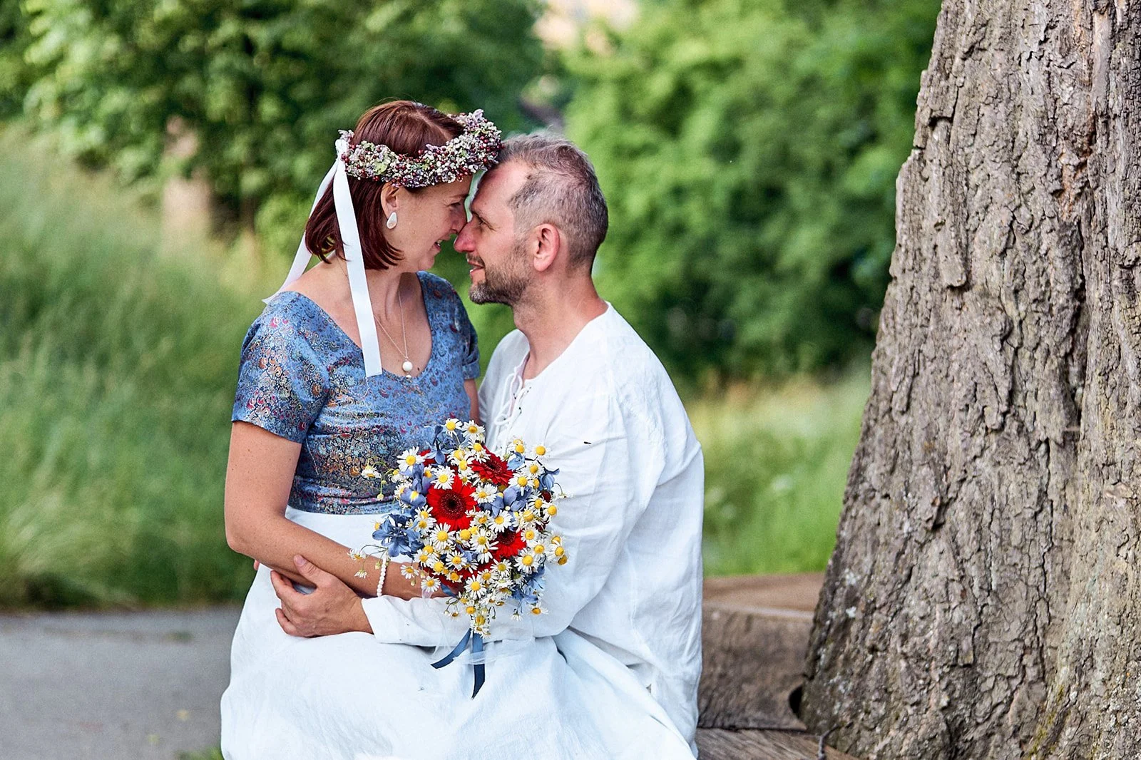 A couple sitting on a park bench, leaning in and touching foreheads, with a large tree trunk to their right. The woman is wearing a blue patterned dress and a floral headband, holding a colorful bouquet of flowers. The man is dressed in a white shirt