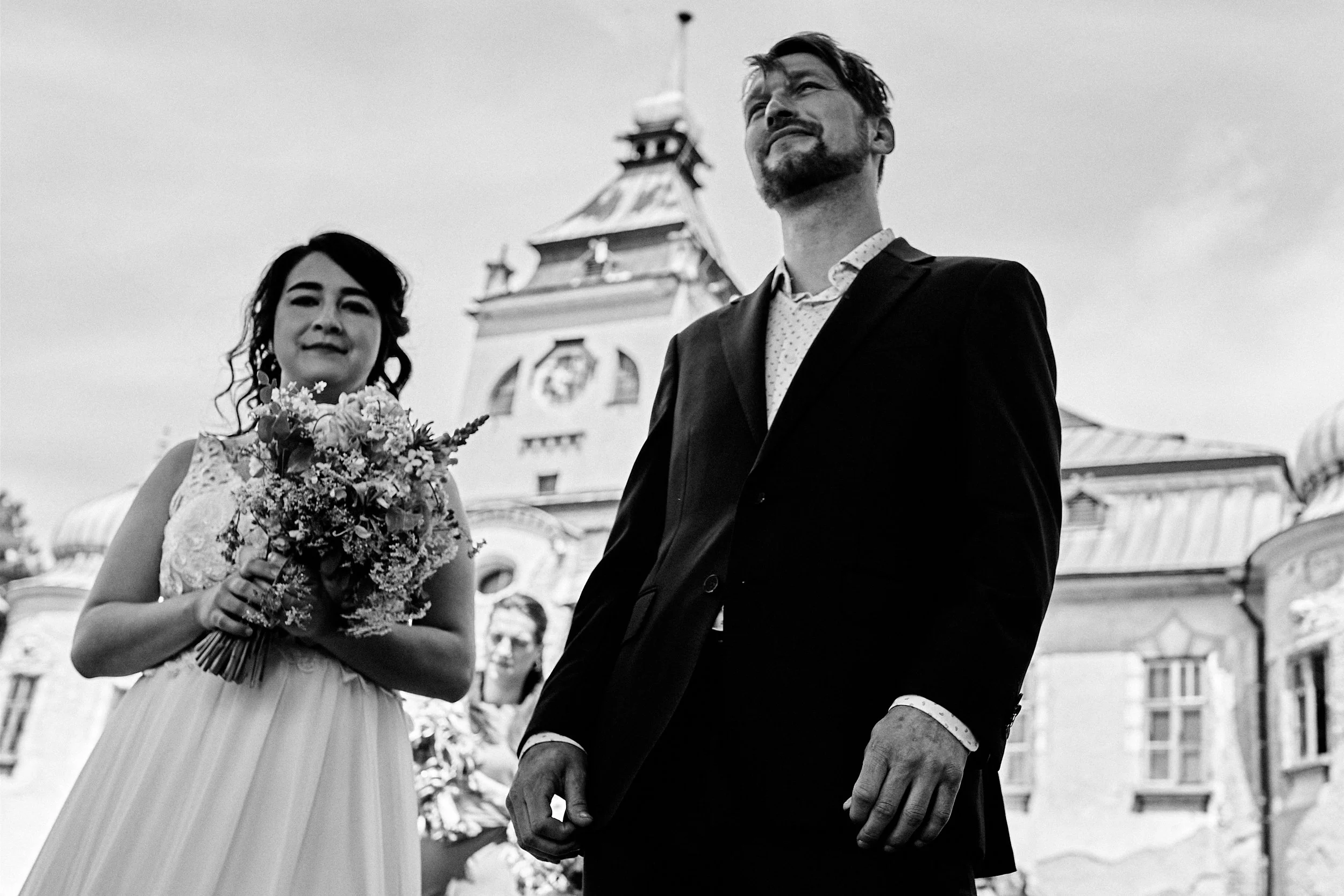 A black-and-white photo of a couple, likely newlyweds, standing outdoors. The woman is holding a bouquet of flowers and smiling softly, while the man, dressed in a suit, is gazing upward with a confident expression. There are people and historic buil