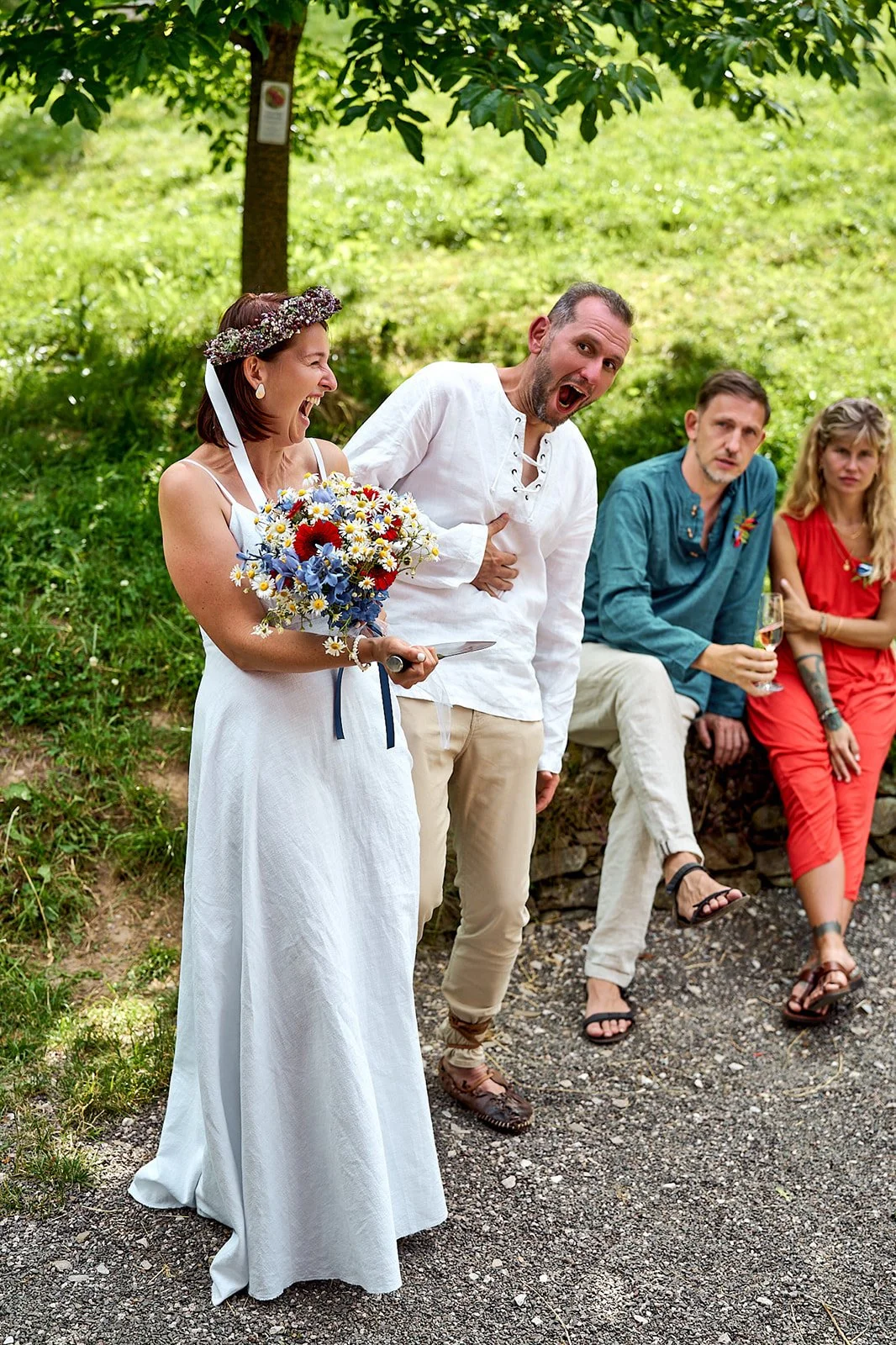People at an outdoor gathering, with a woman in a white dress and flower crown holding a bouquet, smiling and laughing, and a man in a white shirt standing beside her. Three other individuals sit on a stone wall in the background, one holding a glass