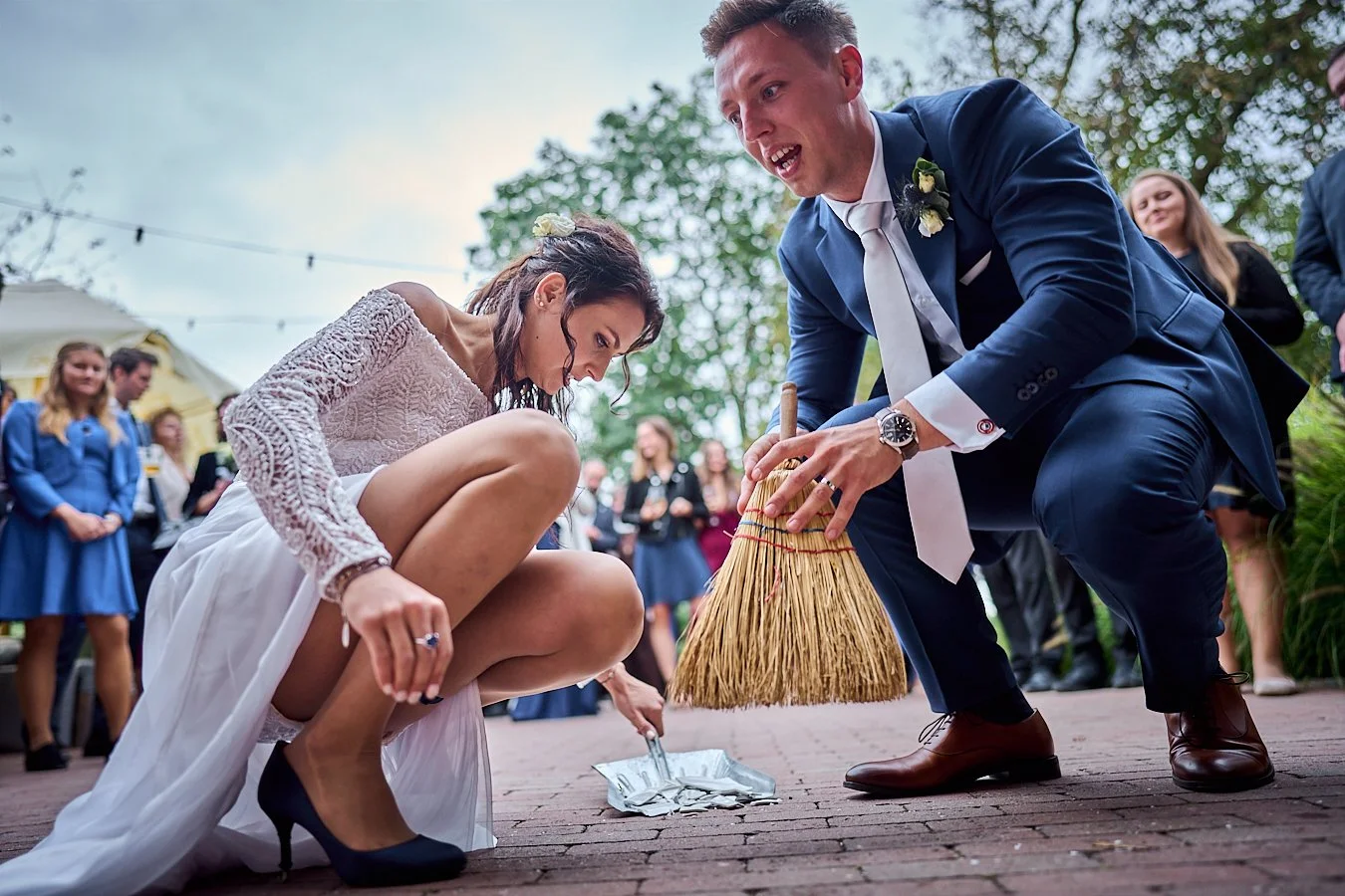 Couple dressed in wedding attire, with the bride crouching and the groom sweeping the ground with a broom during a wedding ceremony outdoors, surrounded by guests.