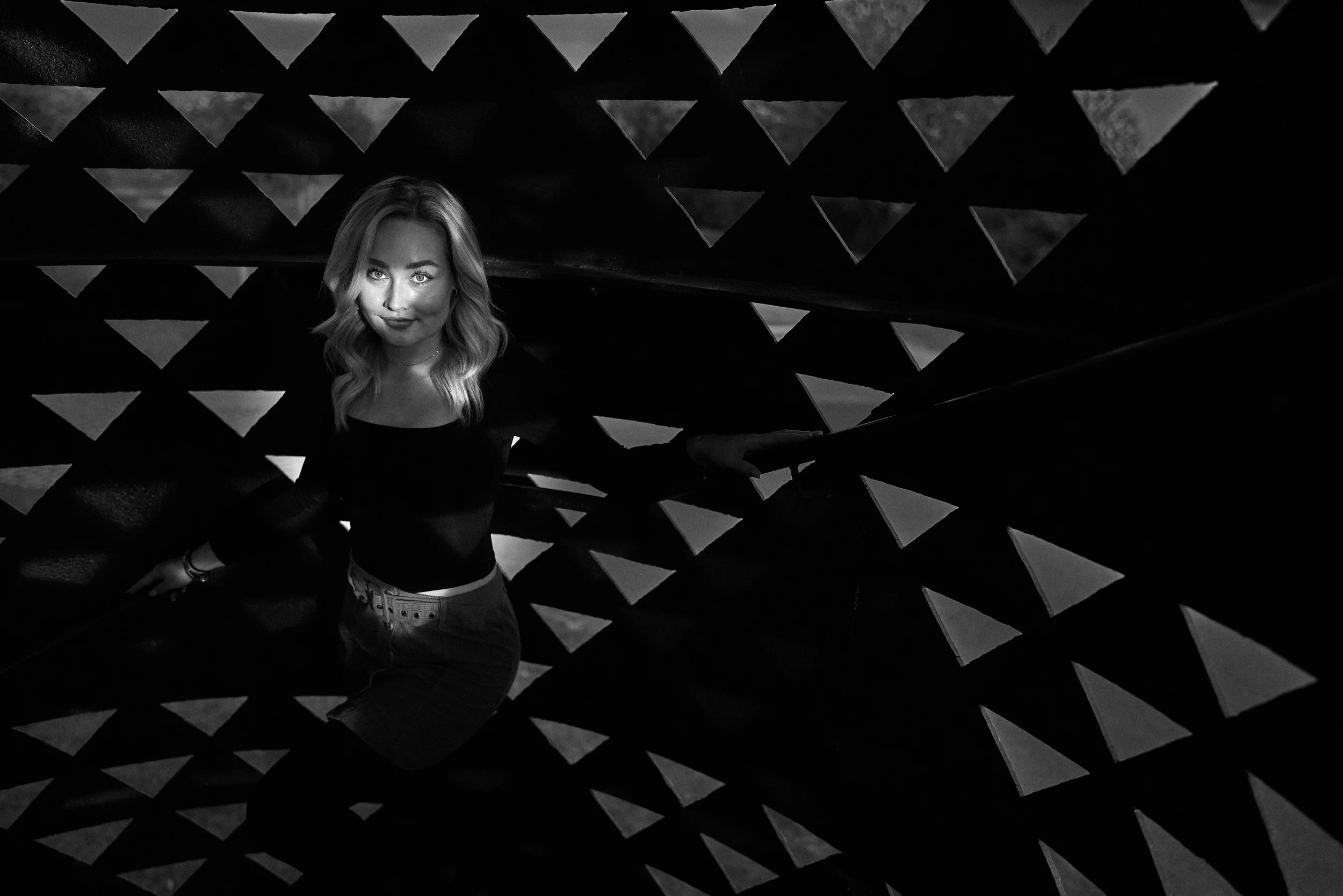 A woman with wavy hair and a confident expression is standing in front of an abstract black and white geometric background with triangle patterns.