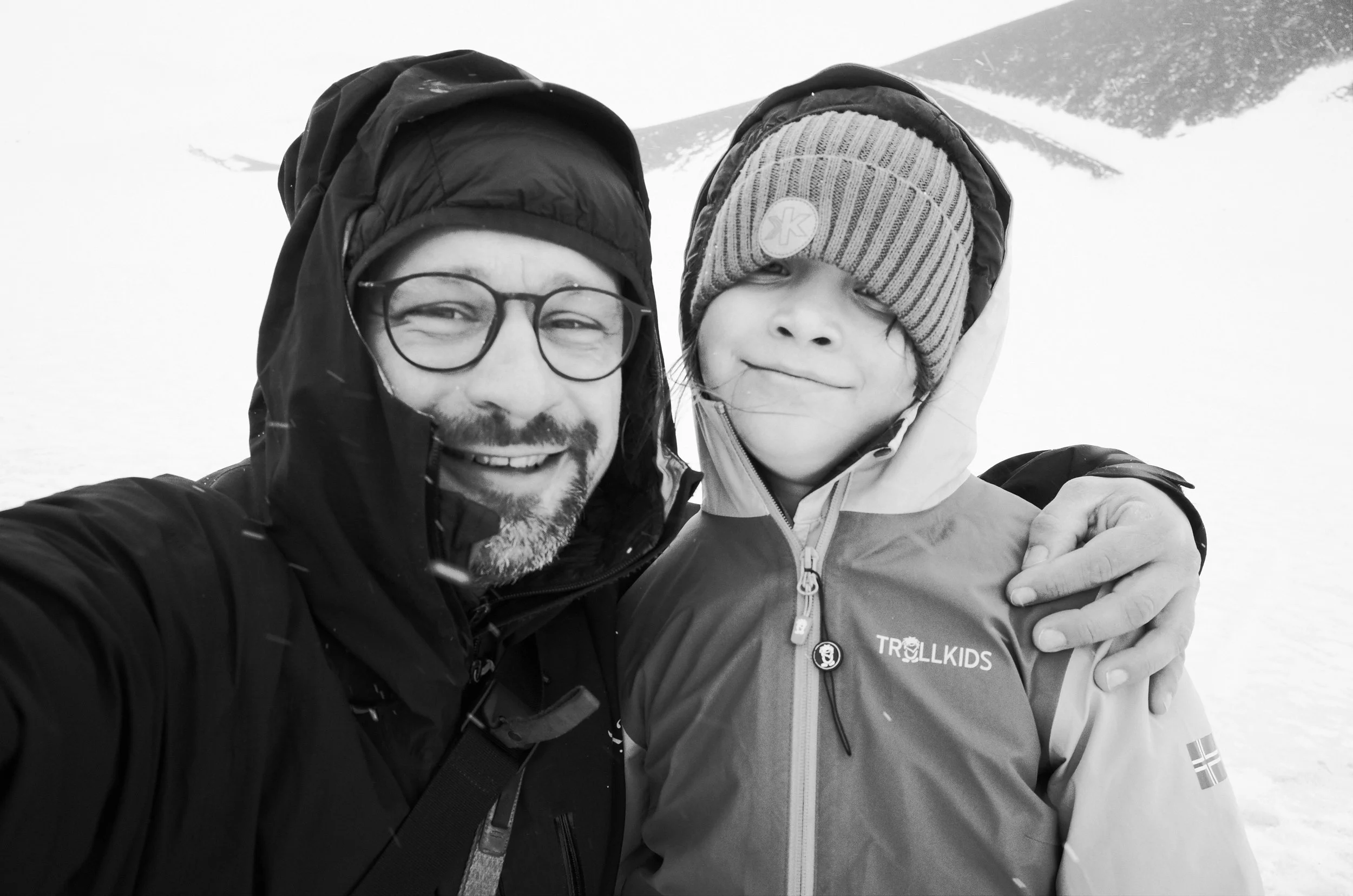 A man with glasses and a beard taking a selfie with a child in snowy outdoors. Both are bundled up in winter jackets and hats, with snow falling around them.