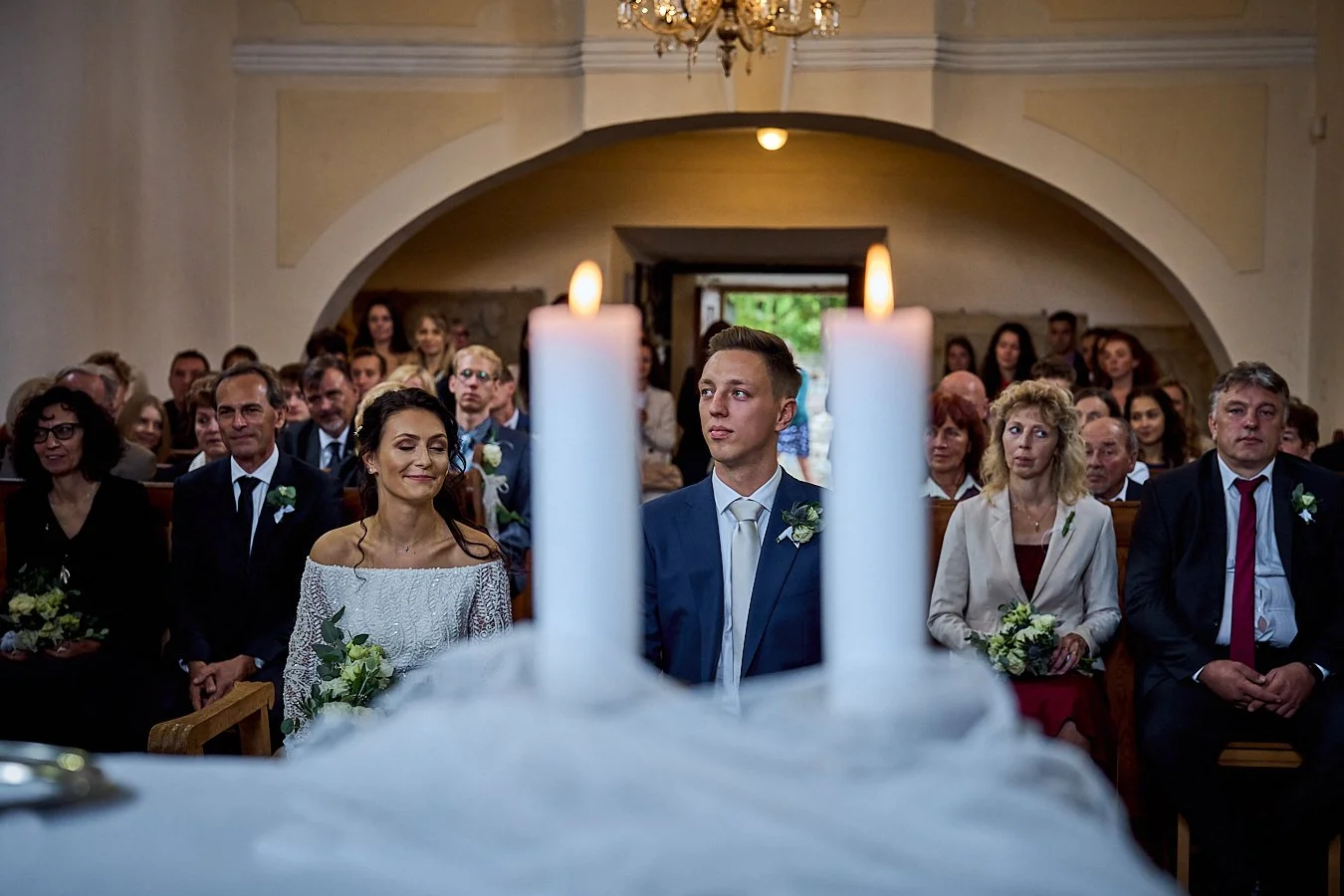 A wedding ceremony inside a church, with the bride and groom sitting in the front row, facing the altar with lit candles, surrounded by family and friends.