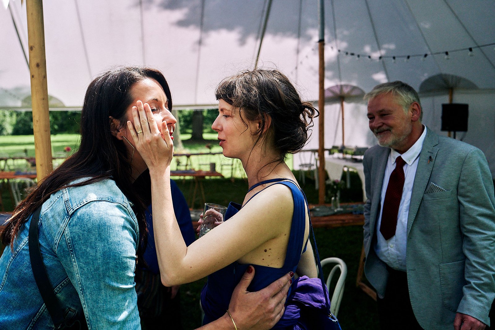 A woman with long dark hair in a denim jacket is crying while another woman in a blue dress comforts her at an outdoor event under a large white tent, with a man in a grey suit smiling nearby.