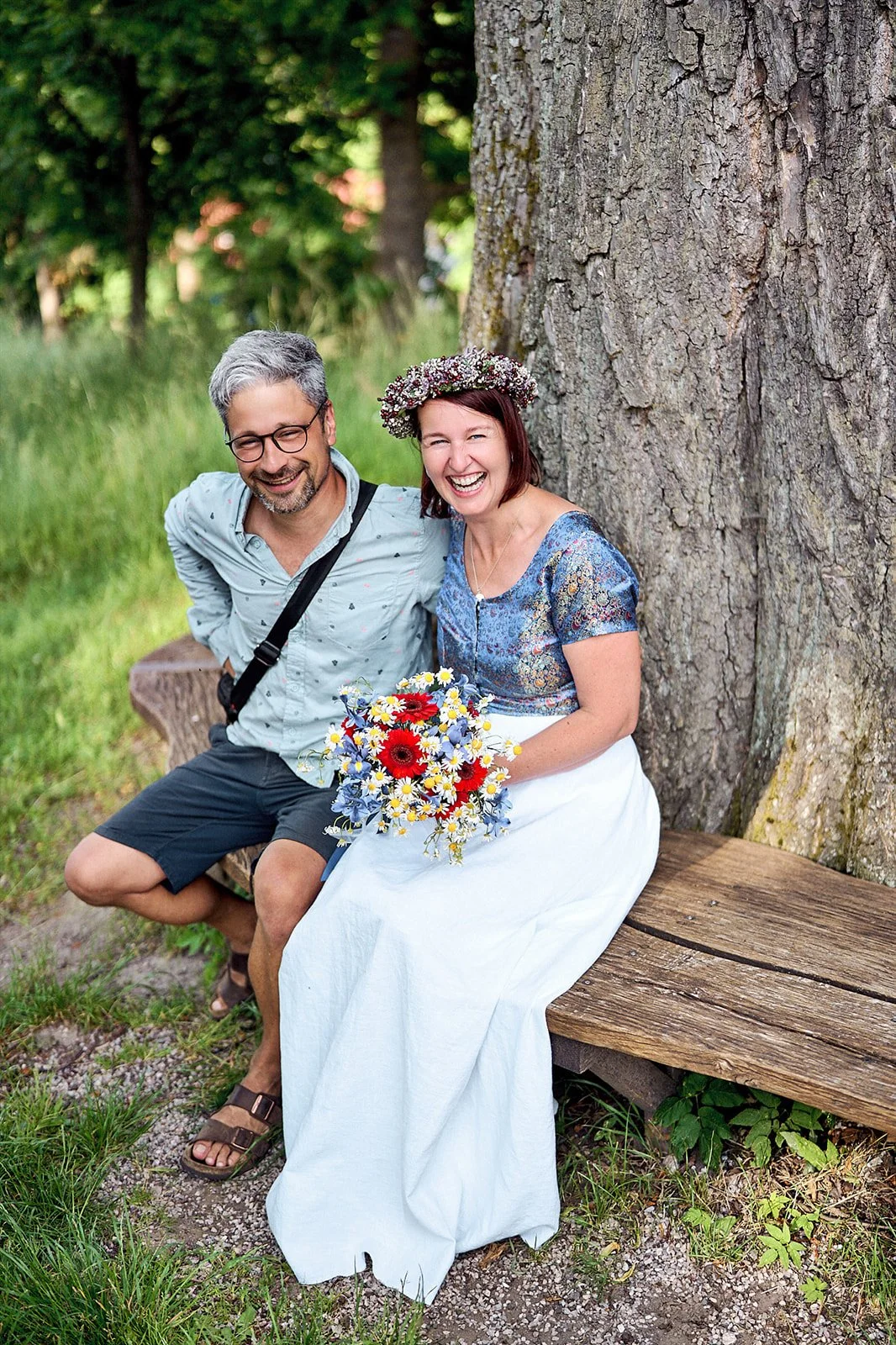 A happy couple sitting on a wooden bench outdoors beside a large tree. The woman is wearing a floral crown and holding a bouquet of colorful flowers, and the man is wearing glasses, a light-colored shirt, and shorts. They are smiling and enjoying the moment.