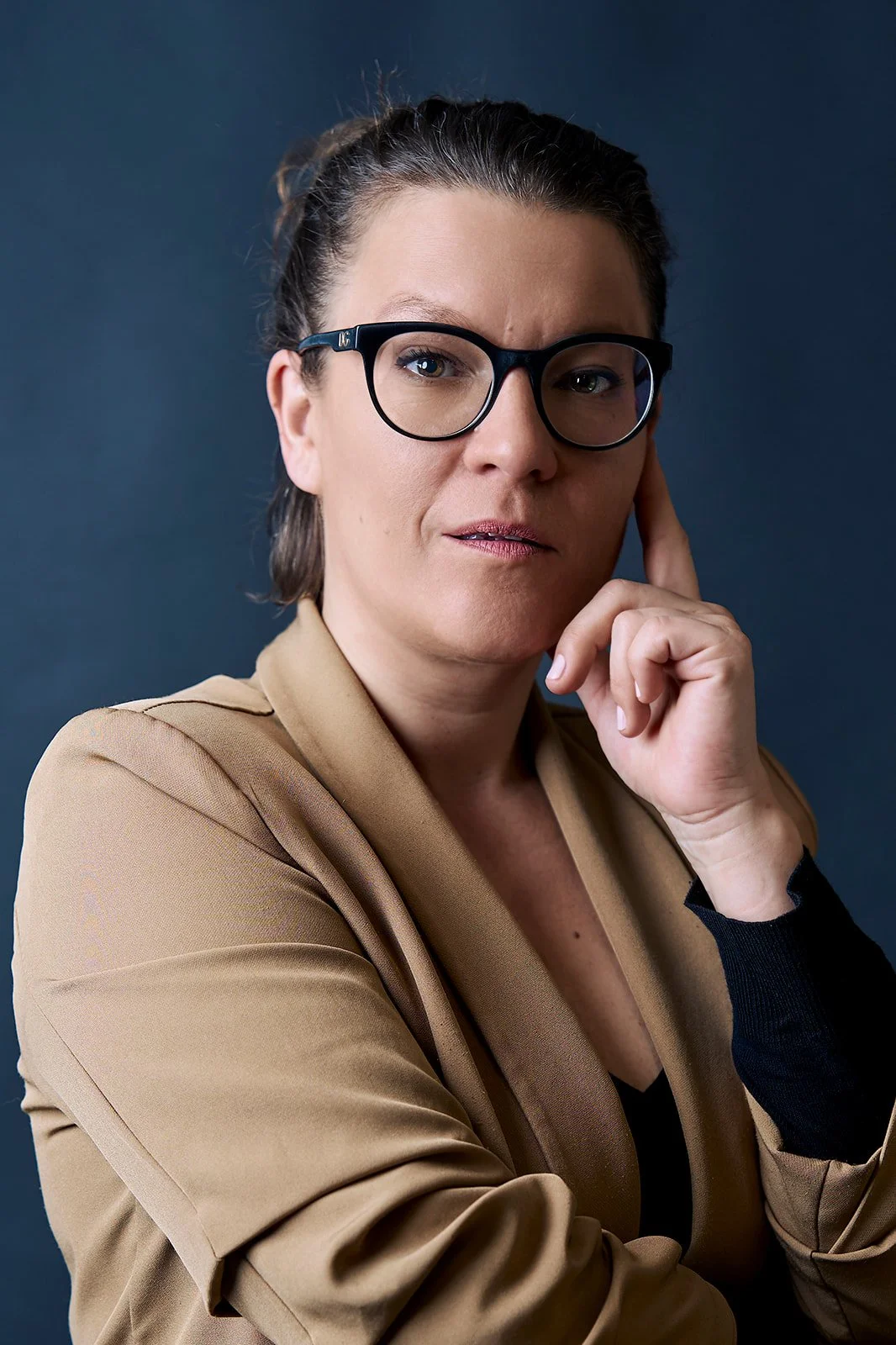 A woman wearing glasses and a beige blazer, with her hand on her cheek, looking thoughtfully at the camera against a dark background.