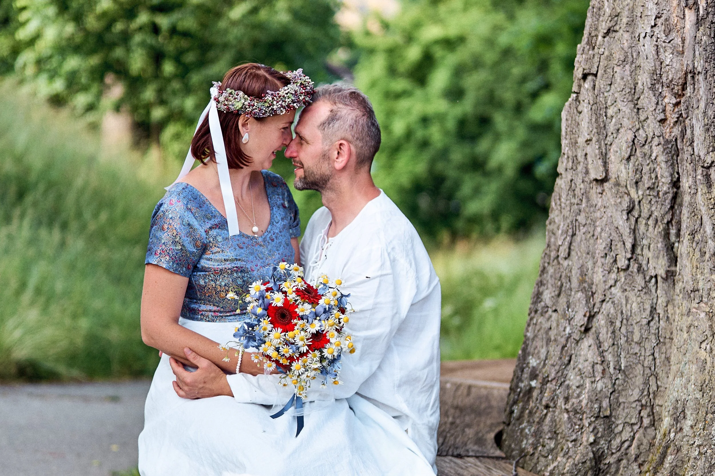 A couple faces each other with foreheads touching, standing outdoors near a large tree, holding a bouquet of colorful flowers, and smiling warmly.