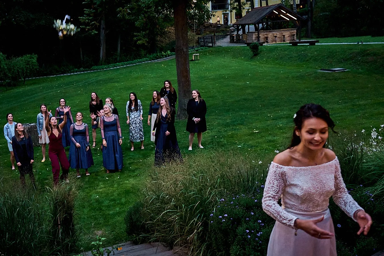 A woman in a white dress stands in the foreground smiling, while a group of women in formal dresses gather on the lawn behind her, enjoying an outdoor event or wedding reception during twilight.