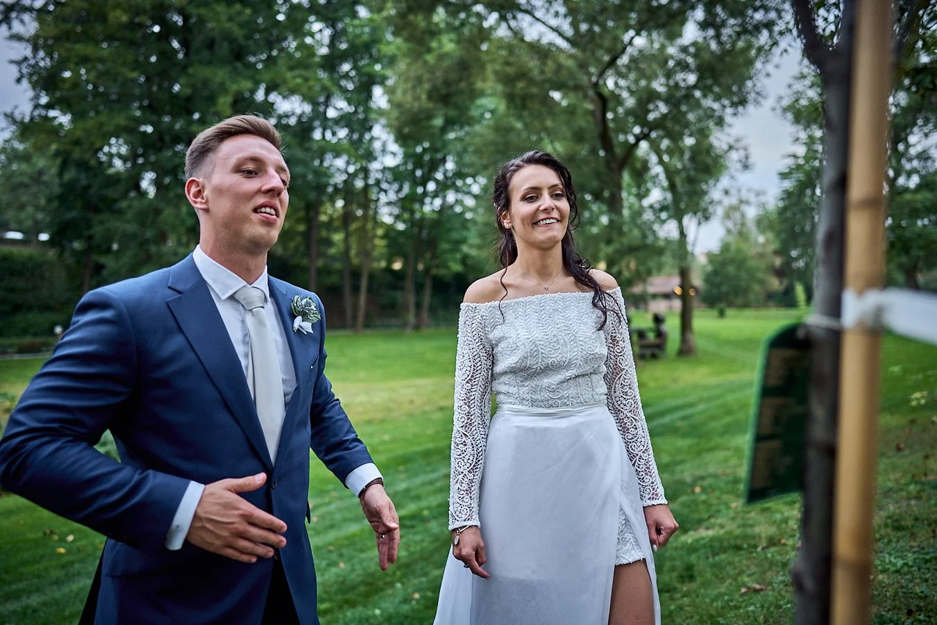 A bride and groom standing outdoors in a park during their wedding, smiling and looking at the camera, with trees and grass in the background.