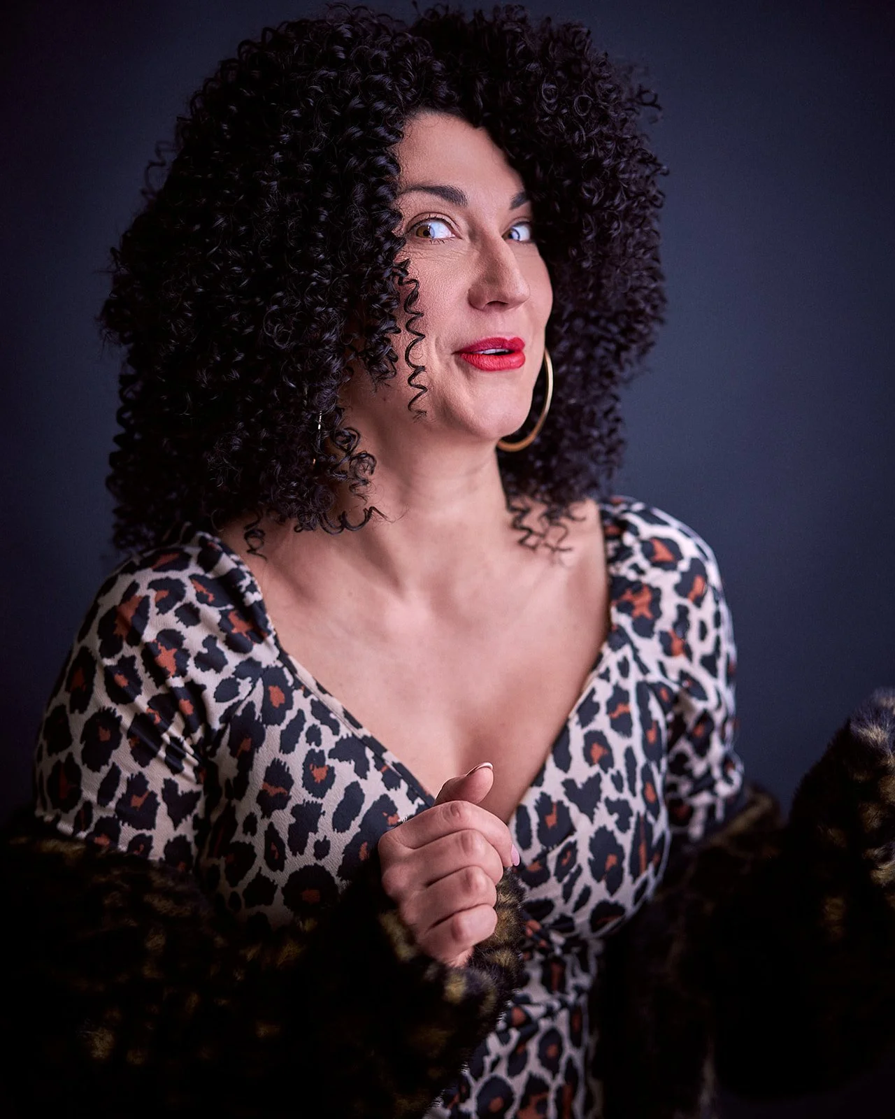 A woman with dark curly hair, red lipstick, wearing a leopard print top, large hoop earrings, and a mink fur coat, posing against a dark background.