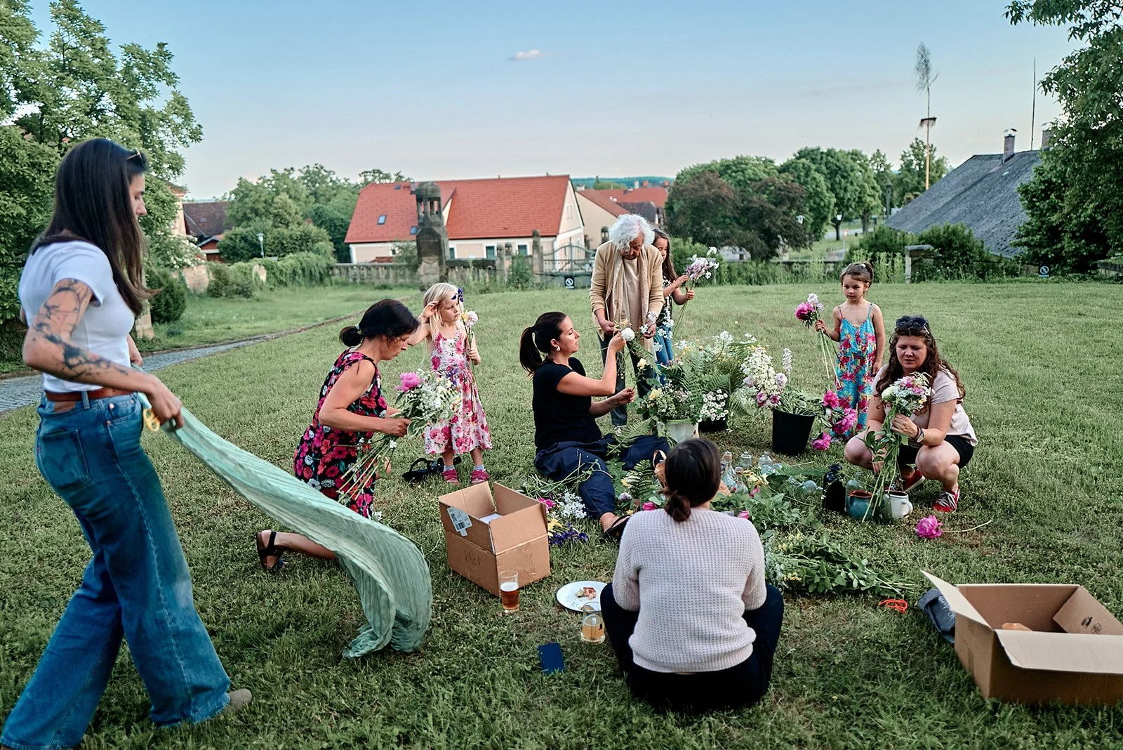 A group of women and children outdoors creating flower arrangements on a grassy lawn with houses and trees in the background.