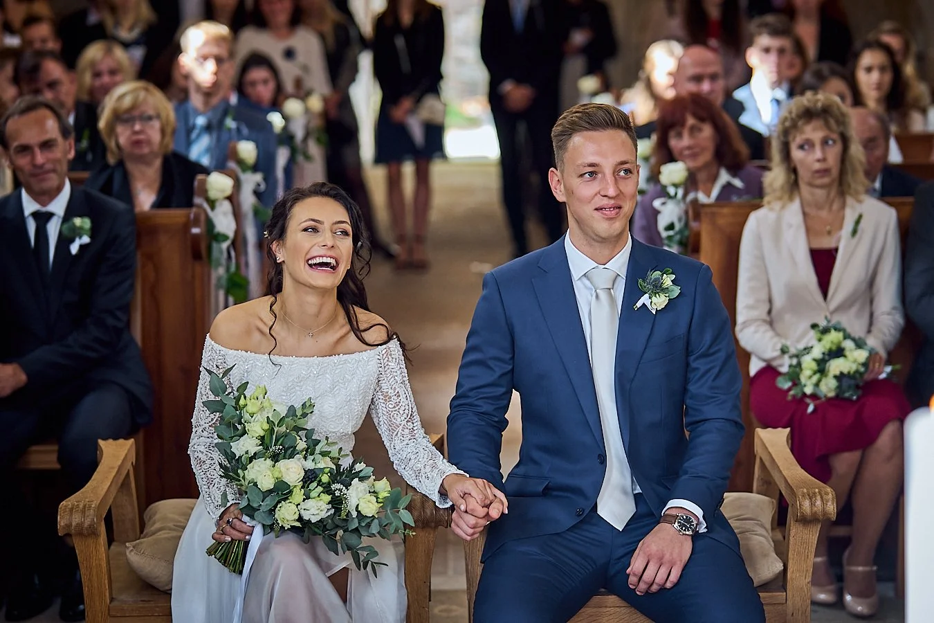 A bride and groom sitting at the altar during their wedding ceremony, holding hands and smiling. The bride wears a white lace dress and holds a bouquet of white and green flowers, while the groom is dressed in a blue suit with a white tie and boutonn