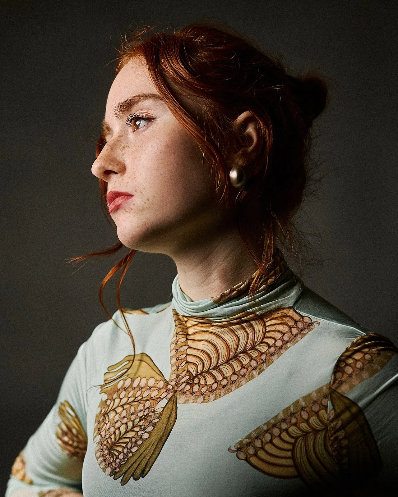 Side profile of a woman with red hair, freckles, and pearl earring, wearing a white top with butterfly and moth designs.