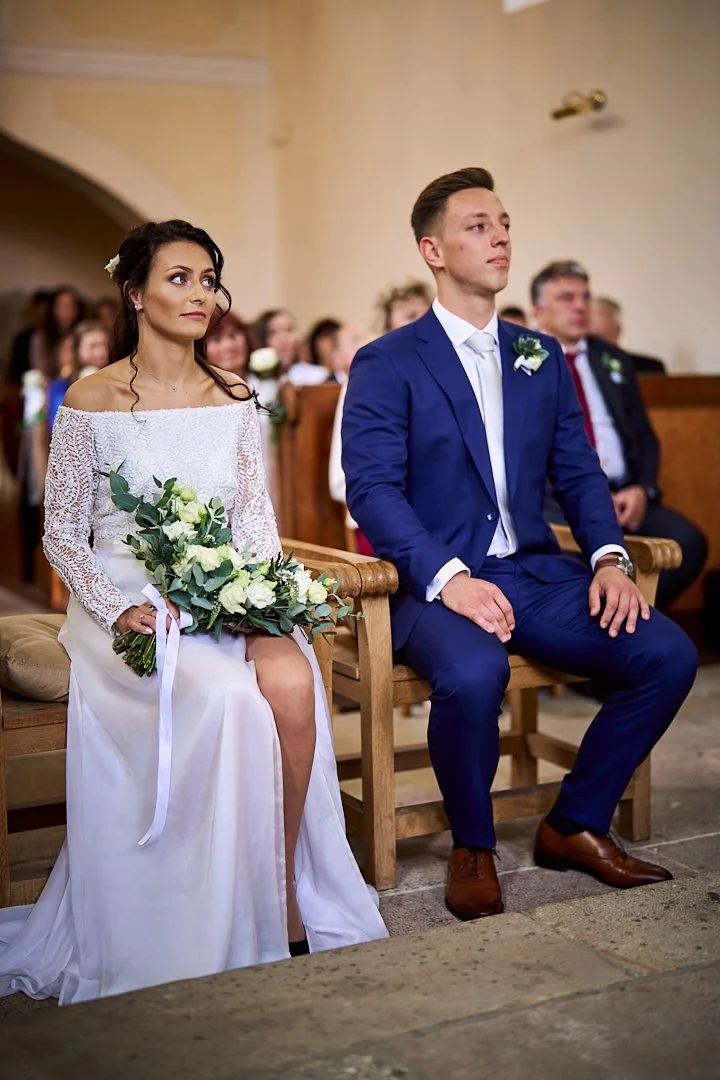 A bride and groom sitting in a church during a wedding ceremony, with bridesmaids and guests visible in the background.
