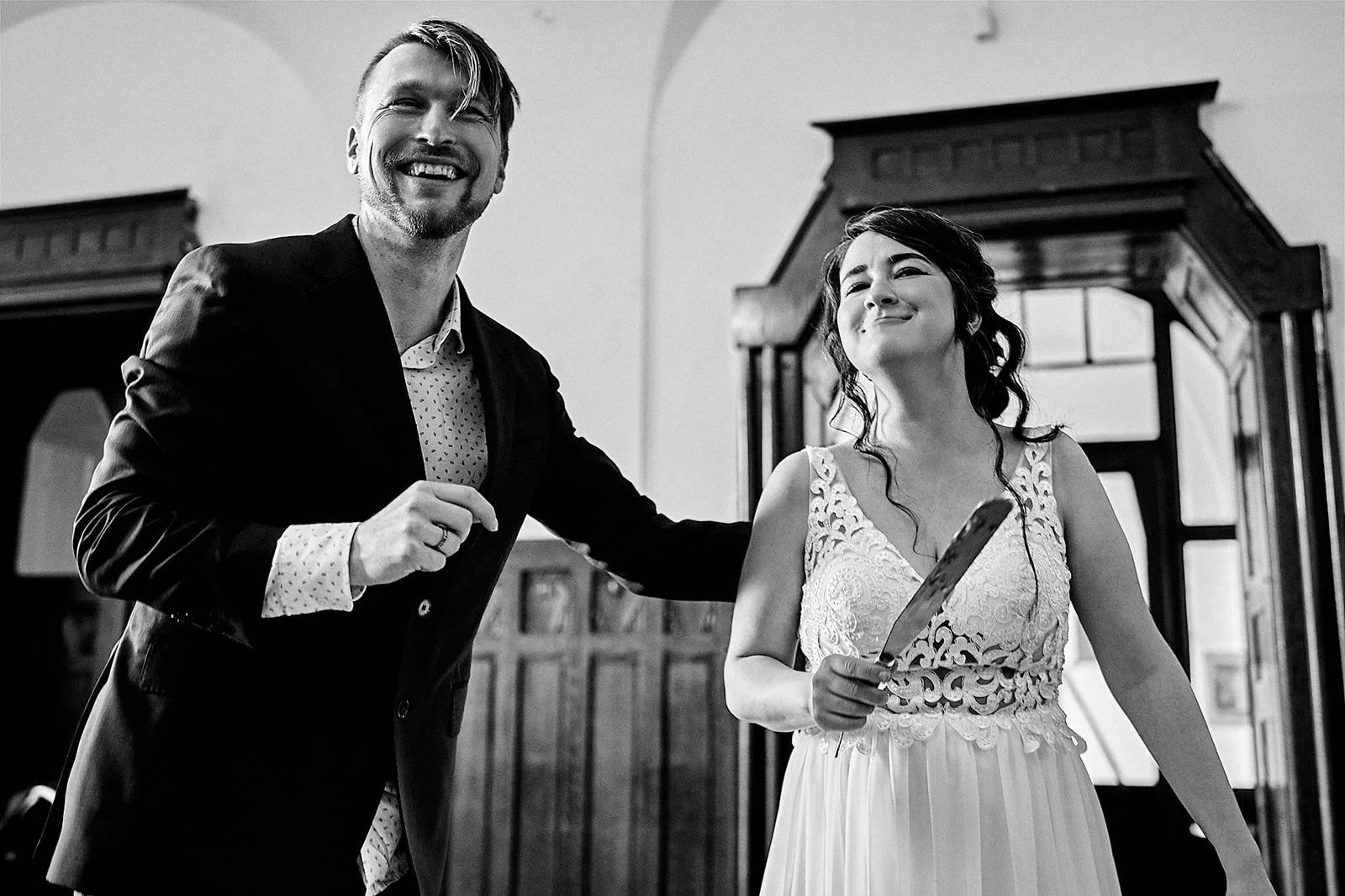A black and white photo of a man and woman smiling. The woman is holding a knife and wearing a lace dress, while the man is wearing a suit with a patterned shirt. They are in a room with wooden furniture and decorative elements.