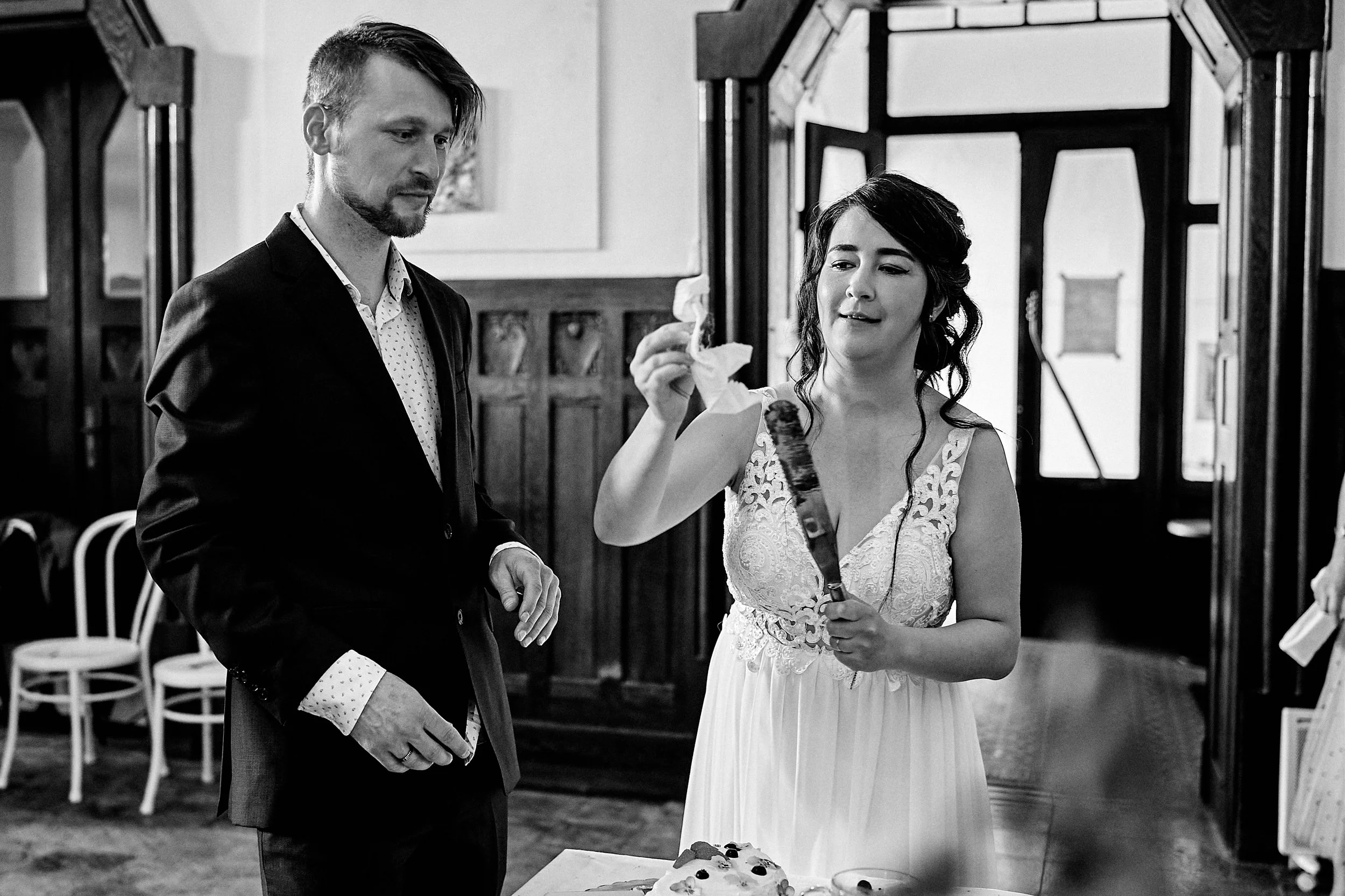 A black and white photo of a bride and groom at their wedding reception. The groom in a suit and polka dot shirt is looking at a wedding cake, while the bride in a lace wedding dress is holding a knife, about to cut the cake. There are chairs and a d