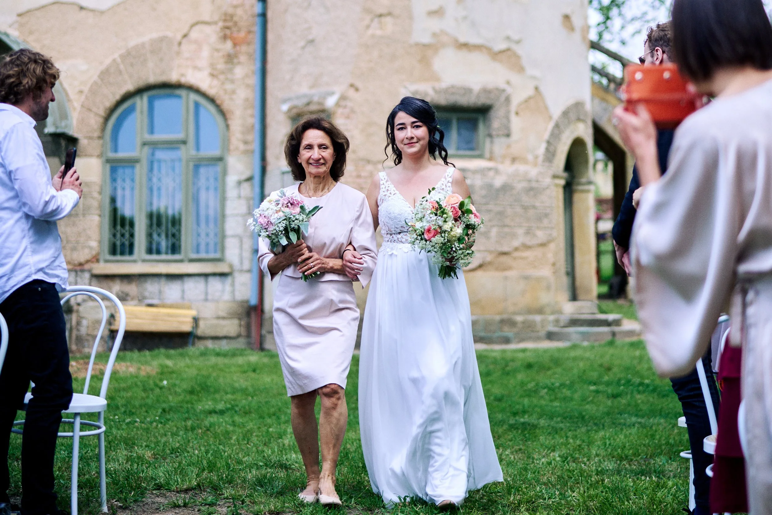 A bride in a white wedding gown walking down the aisle with an older woman, possibly her mother, holding a bouquet of flowers. The ceremony appears to be outdoors in front of an old stone building, with guests seated on white chairs.