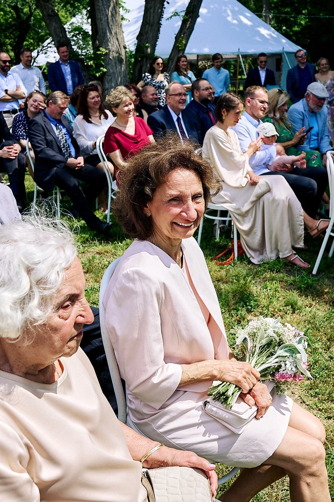People attending an outdoor wedding ceremony, seated on white chairs, with a large white tent and trees in the background. The woman in the foreground is holding a bouquet of flowers and smiling.