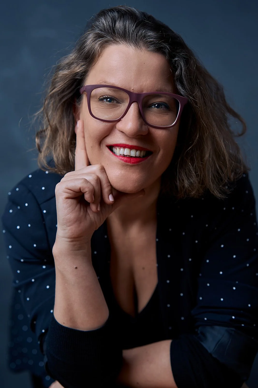 A woman with glasses, smiling, resting her chin on her hand, wearing a dark shirt with white polka dots.