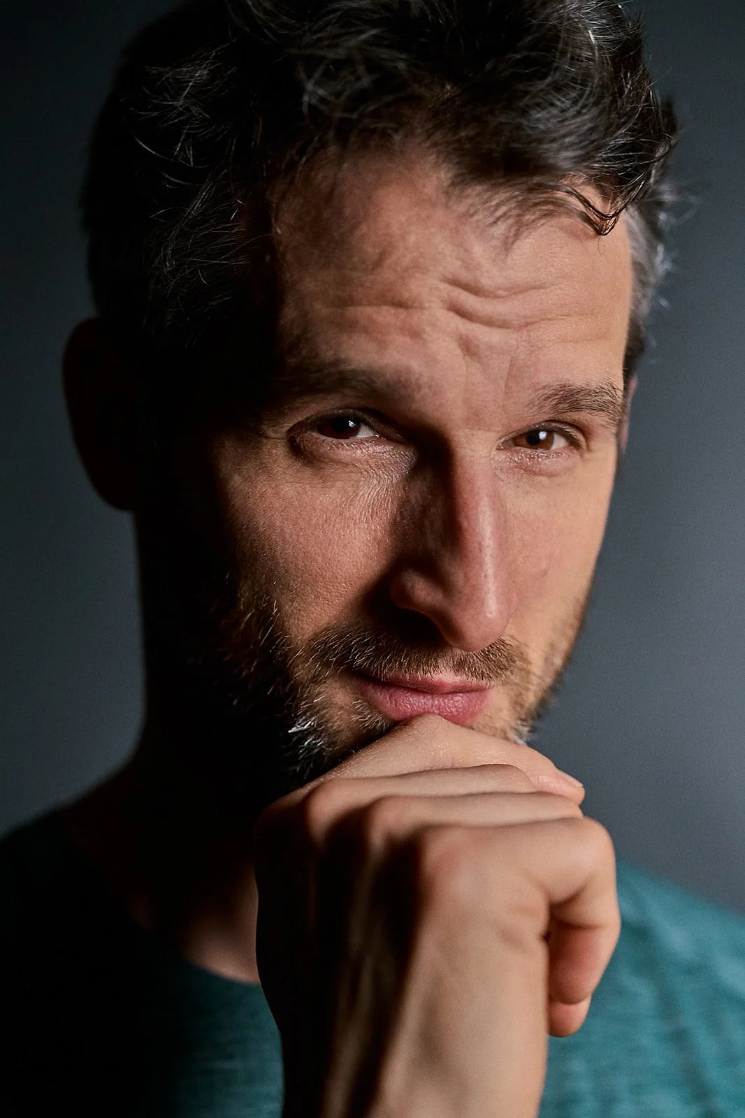 Close-up of a man with brown hair and beard, resting his chin on his hand, looking at the camera with a contemplative expression.
