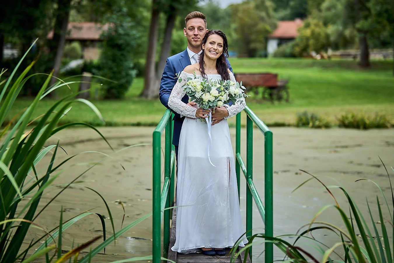 A bride and groom standing on a small dock over a pond, with the bride holding a bouquet of white and green flowers. The groom is in a blue suit, and they are smiling. There are trees and a house in the background.