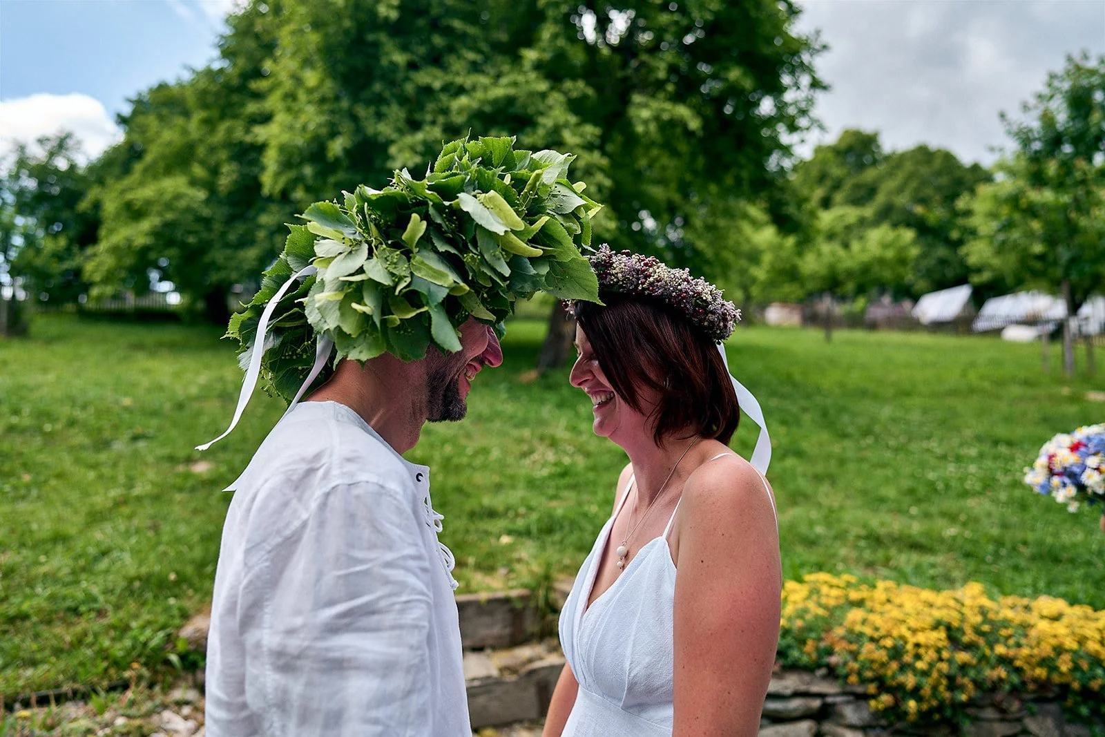 A couple smiling at each other outdoors, wearing flower crowns with greenery and flowers, in a park setting with trees and flowers in the background.