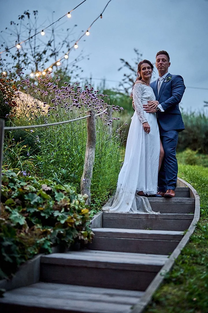 A couple dressed in wedding attire standing on wooden steps outdoors during evening. The bride is wearing a white lace dress with long sleeves and a slit, the groom is in a blue suit with a white shirt and blue tie. String lights hung overhead create
