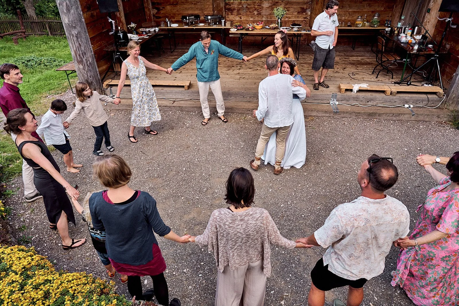 A group of people holding hands and dancing in a circle outdoors at a gathering or celebration, with a rustic wooden stage and buffet table in the background.