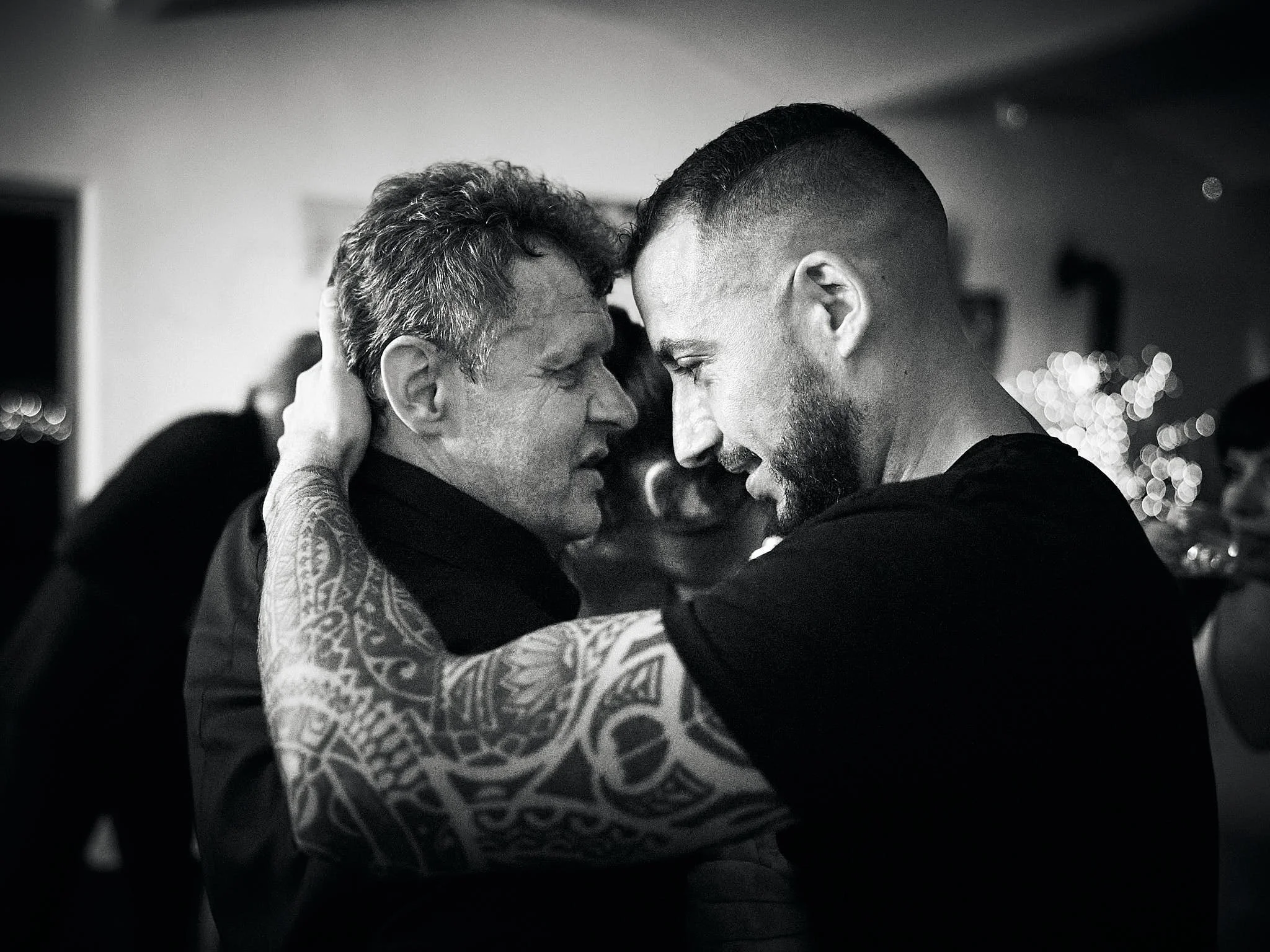 Two men face each other closely, touching foreheads, in an intimate moment at a gathering. The man on the left has curly hair and visible tattoos on his arm, while the man on the right has a short fade haircut and a beard. The background is blurred w