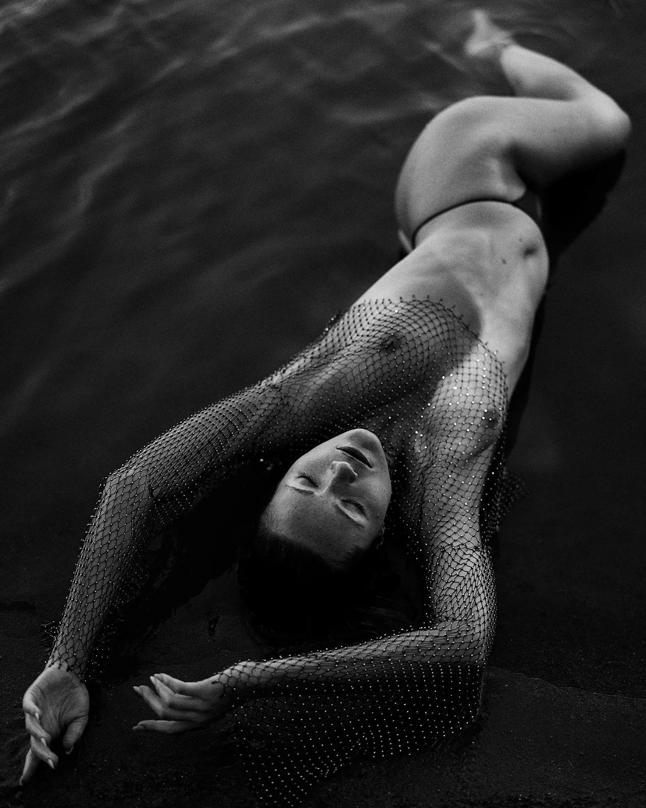 A woman lying on the ground, dressed in a fishnet bodysuit, with her eyes closed and her arms extended above her head.