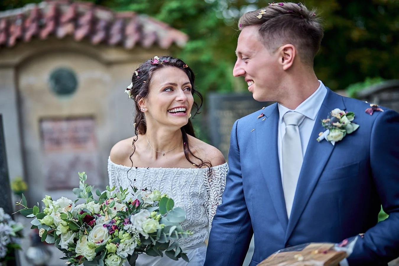 Bride and groom smiling at each other outdoors during wedding, with the bride holding a bouquet of white and purple flowers.