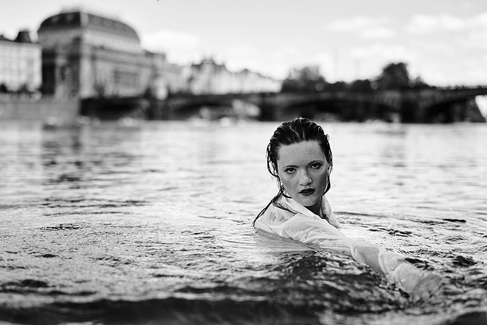 Black and white photo of a woman with wet hair in the water, looking directly at the camera, with a cityscape and bridge in the background.