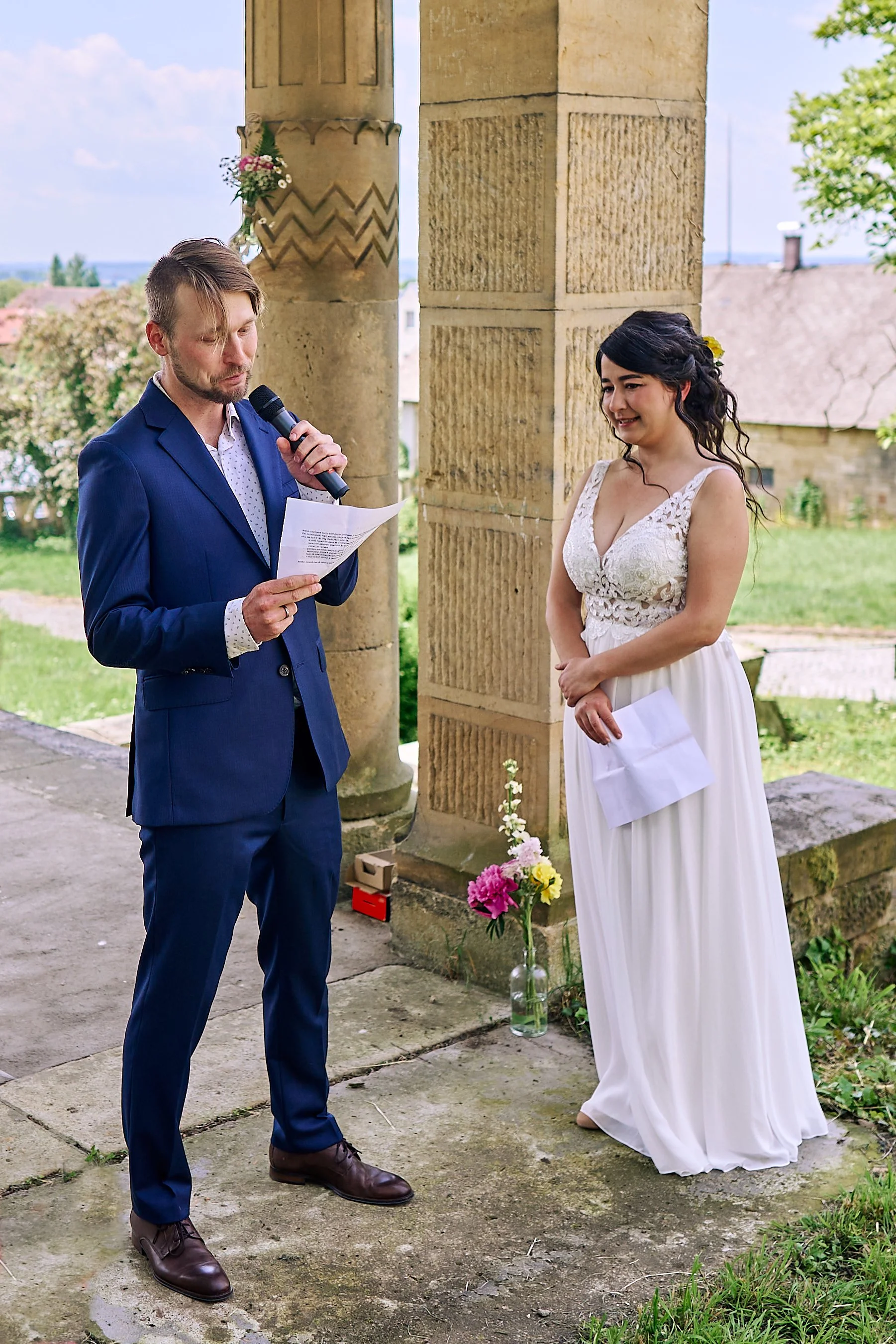 A man in a blue suit and a woman in a white dress are outdoors, with the man reading from a paper and the woman looking at him while holding a piece of paper. There are stone columns, flowers, and a grassy yard in the background.