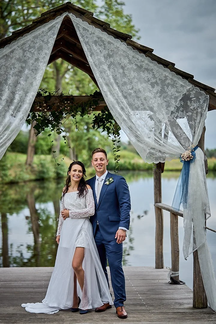 A bride and groom standing on a dock near a lake during their wedding, under a wooden arch decorated with lace curtains and greenery, with trees and water in the background.