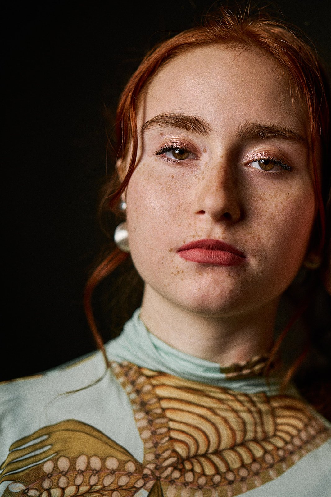 Close-up portrait of a young woman with red hair, freckles, and brown eyes, wearing pearl earrings and makeup, against a dark background.