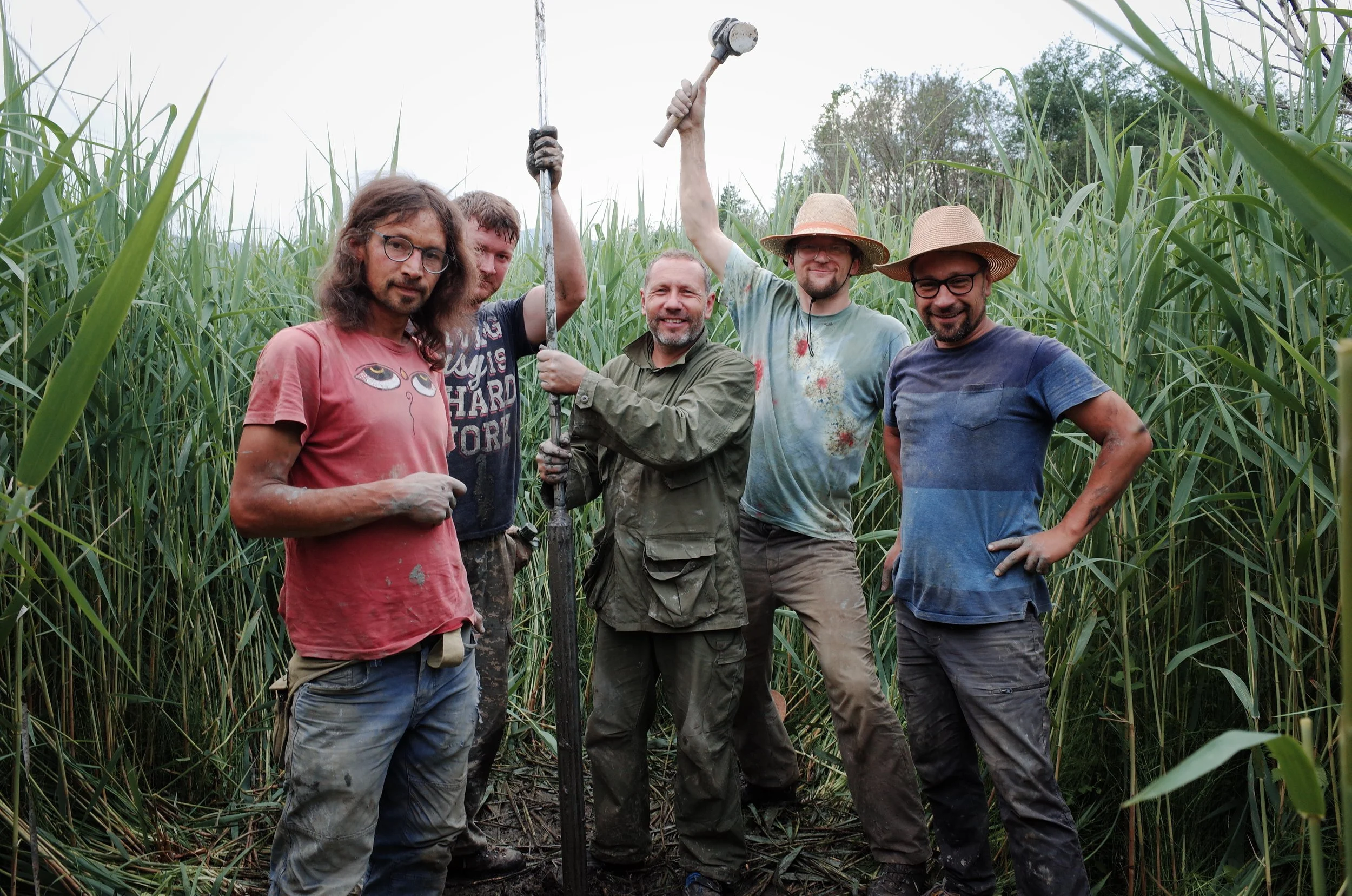 Group of five men standing in a tall grassy field, some holding tools, smiling, with trees in the background.