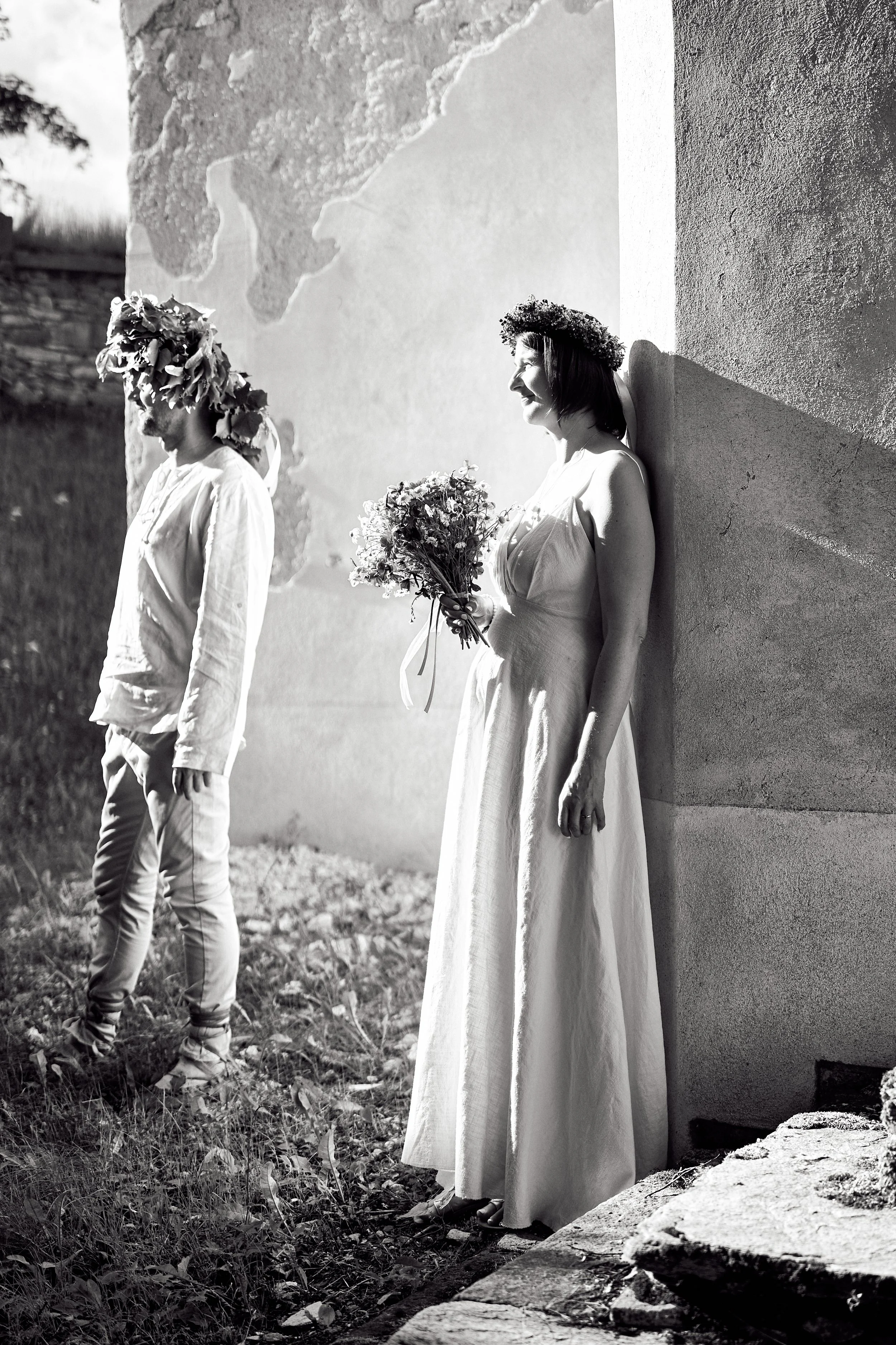 Black and white photo of a woman in a wedding dress and a man in casual clothing, standing outdoors near a wall. The woman is holding a bouquet and wears a floral crown, smiling. The man stands to her left, with a floral headpiece, looking down, with