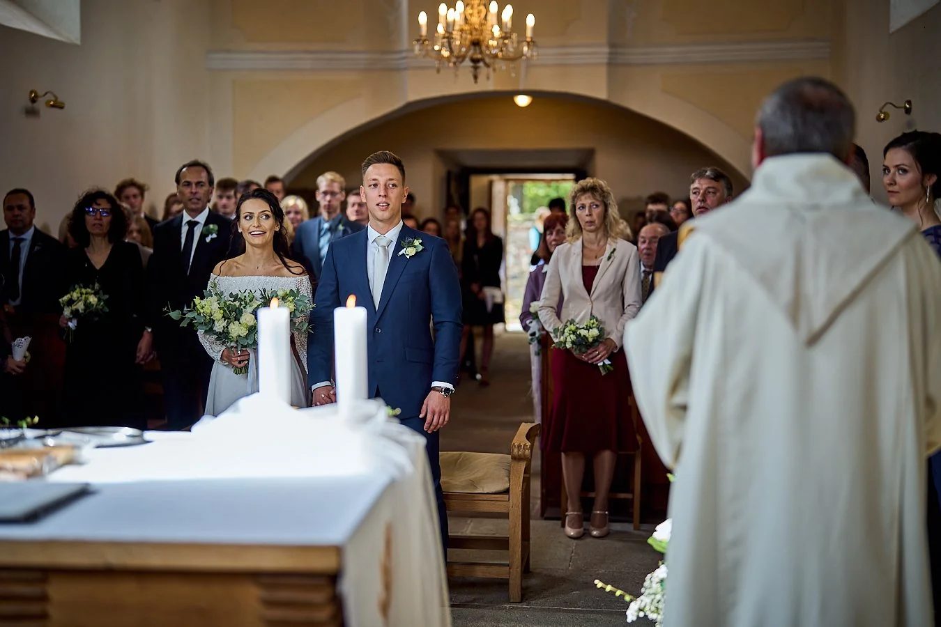 A wedding ceremony with a bride and groom standing in front of a priest, surrounded by guests in a church with a chandelier.