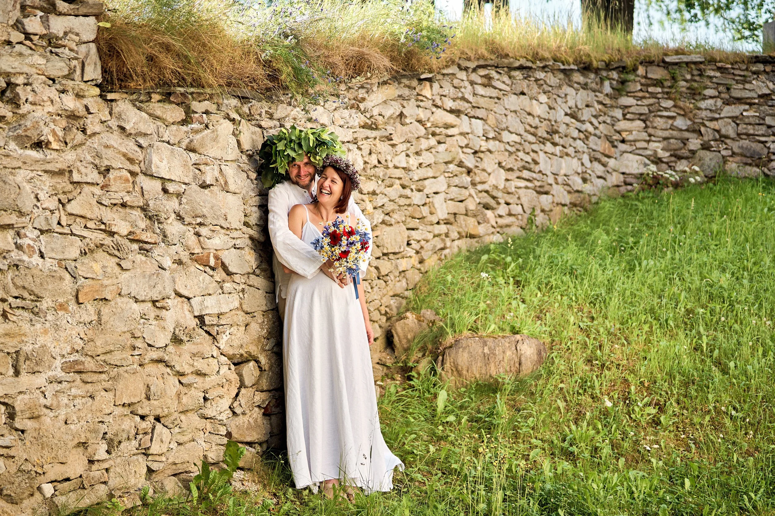 A couple dressed in white, smiling and holding a bouquet of flowers, standing against a stone wall outdoors.