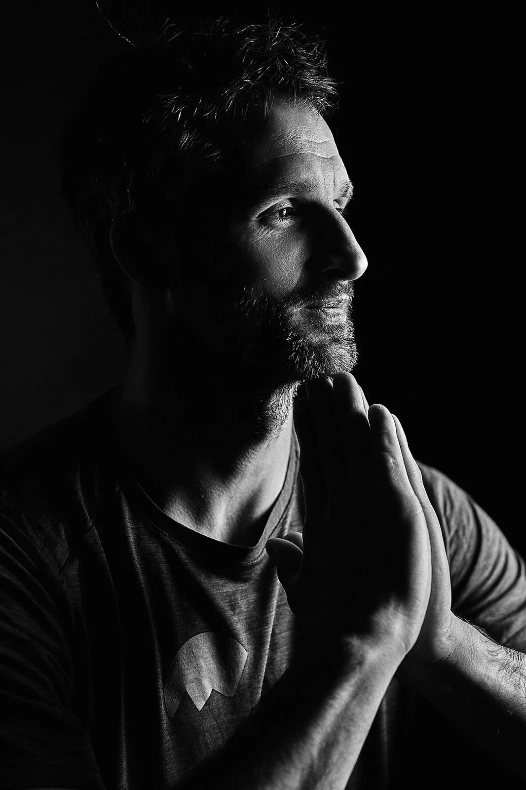 A black and white photo of a man with a beard, his hands pressed together near his face, looking thoughtfully to the side, with dramatic lighting highlighting his profile.