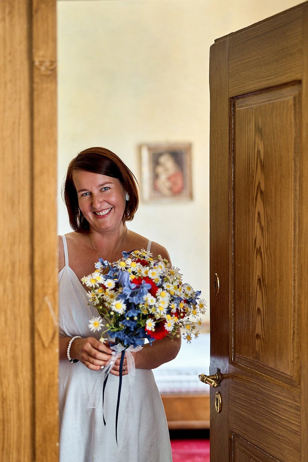A woman with shoulder-length brown hair, wearing a white dress, is smiling while holding a colorful bouquet of flowers.