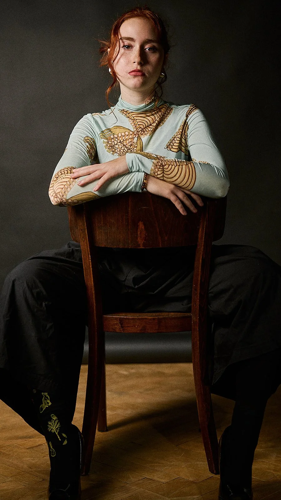 A young woman with red hair sits on a wooden chair against a dark background, wearing a long-sleeved top with butterfly patterns, black pants, and black shoes with embroidered designs. She has her arms crossed on the back of the chair and looks direc
