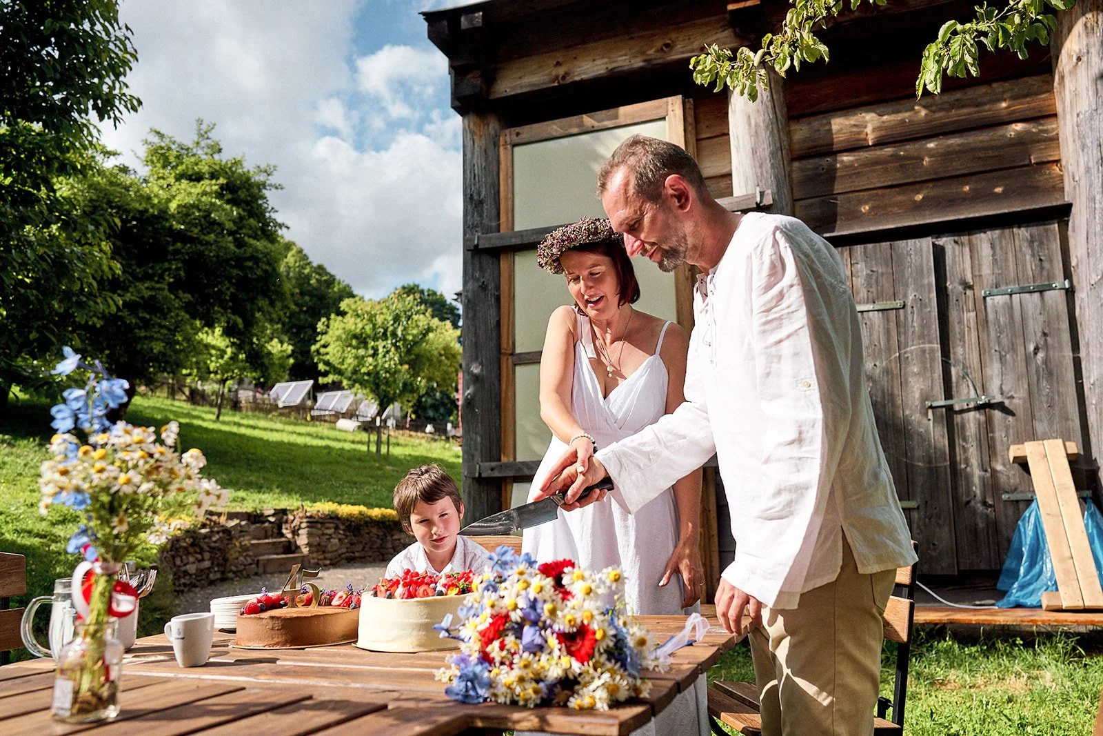 A man, woman, and young boy celebrate outdoors with a cake and flowers, with the man cutting the cake, in front of a rustic wooden building and green trees.
