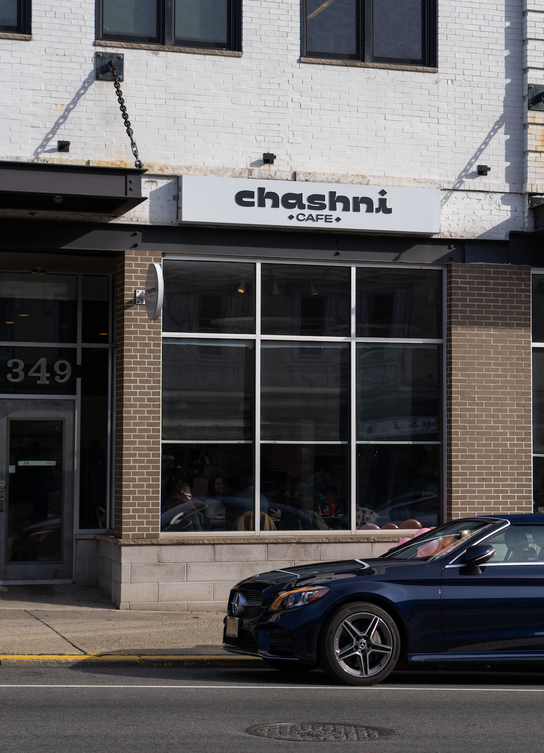 Exterior view of Chashni Cafe with large glass windows, brick and white painted walls, and a black Mercedes-Benz convertible parked in front.