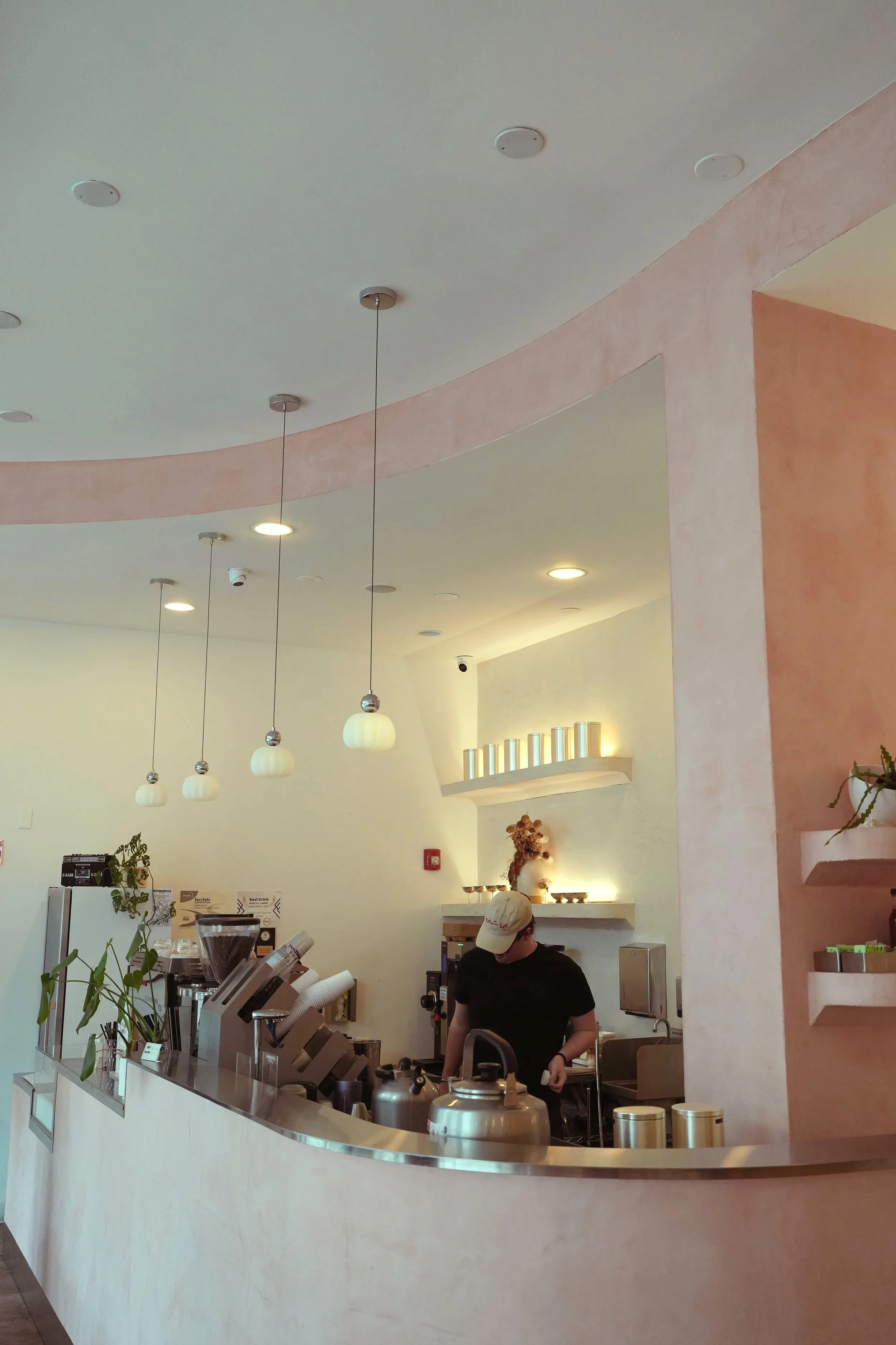 Interior view of a modern coffee shop with a curved white counter, hanging pendant lights, and a person working behind the counter. The background features a pastel pink and beige wall with lighting accents and decorative elements.