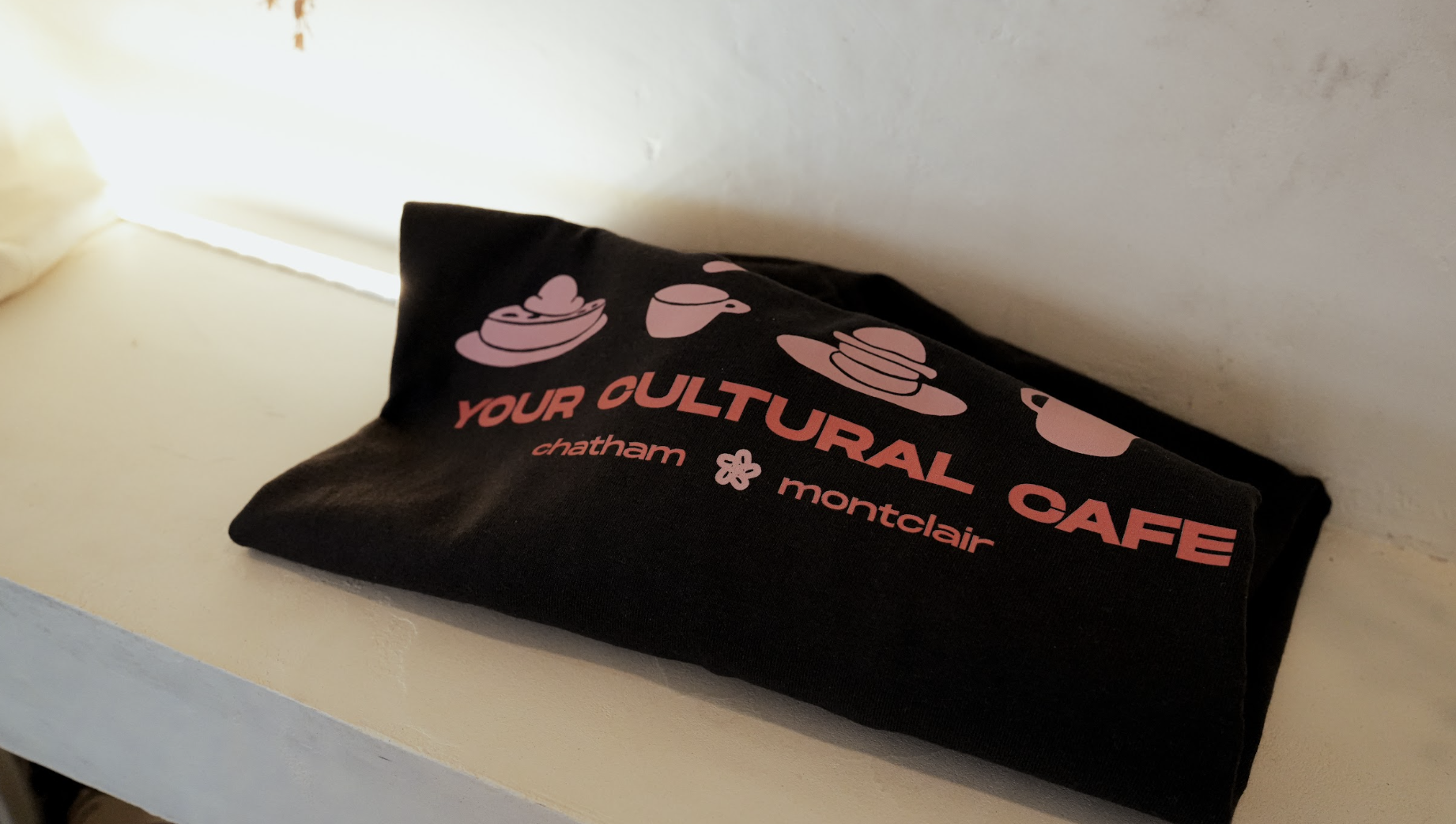 Black fabric item with pink and red text and graphics, reading "Your Cultural Cafe" and listing locations "Chatham" and "Montclair." Includes icons of stacked dishes and a coffee cup, resting on a white surface against a beige wall.