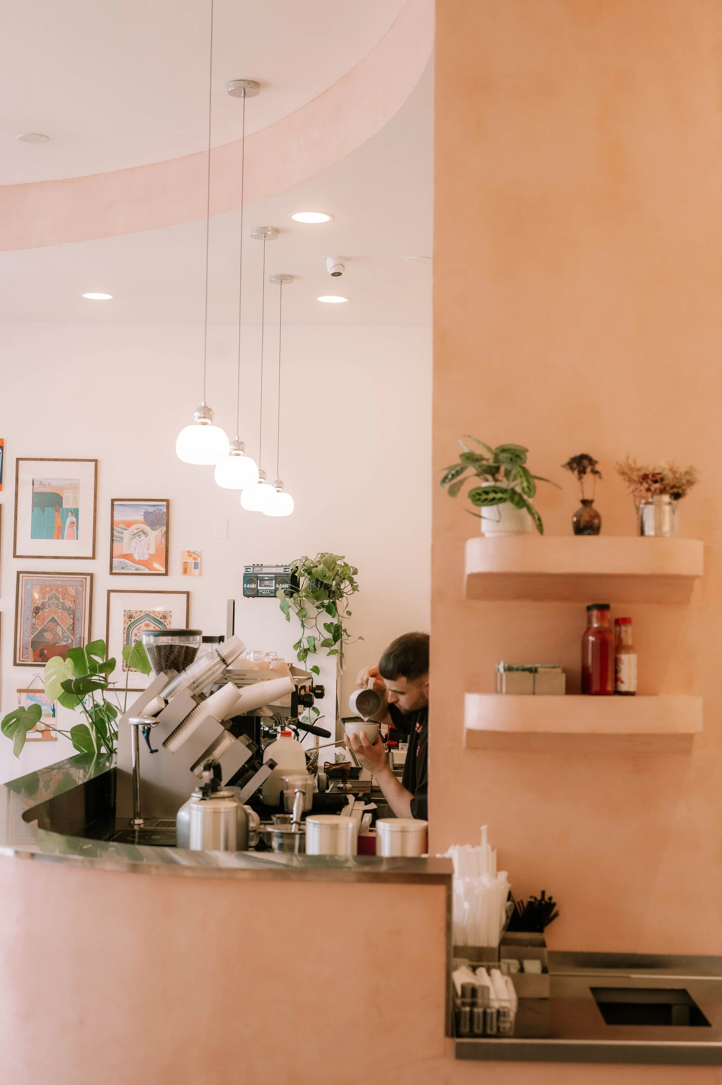 A barista preparing coffee behind the counter of a café, with a pink wall and hanging white pendant lights, and framed artwork on the wall.
