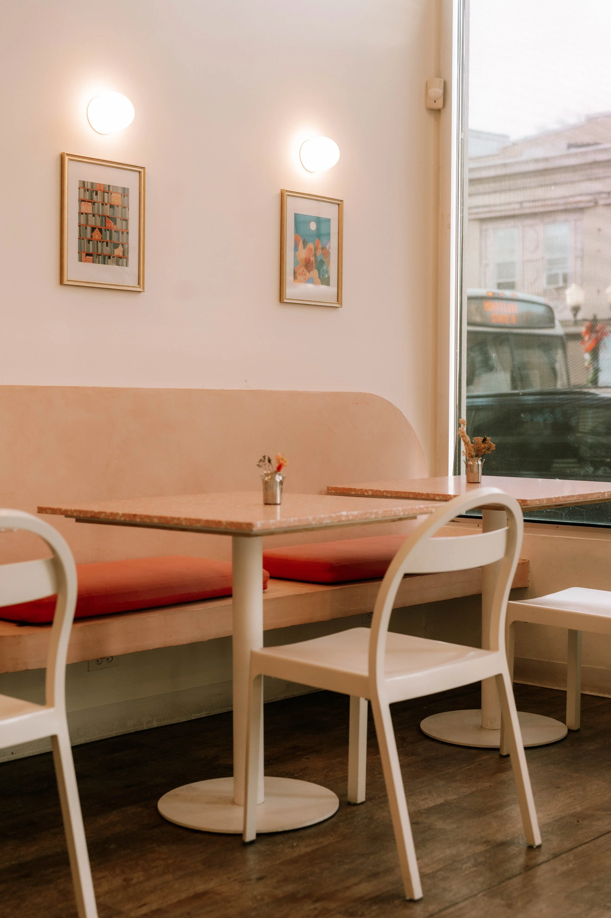 Interior of a cozy cafe with white chairs, pink cushioned seating, a pink table, small vases with flowers, framed artwork on the walls, and a large window looking out onto a city street.