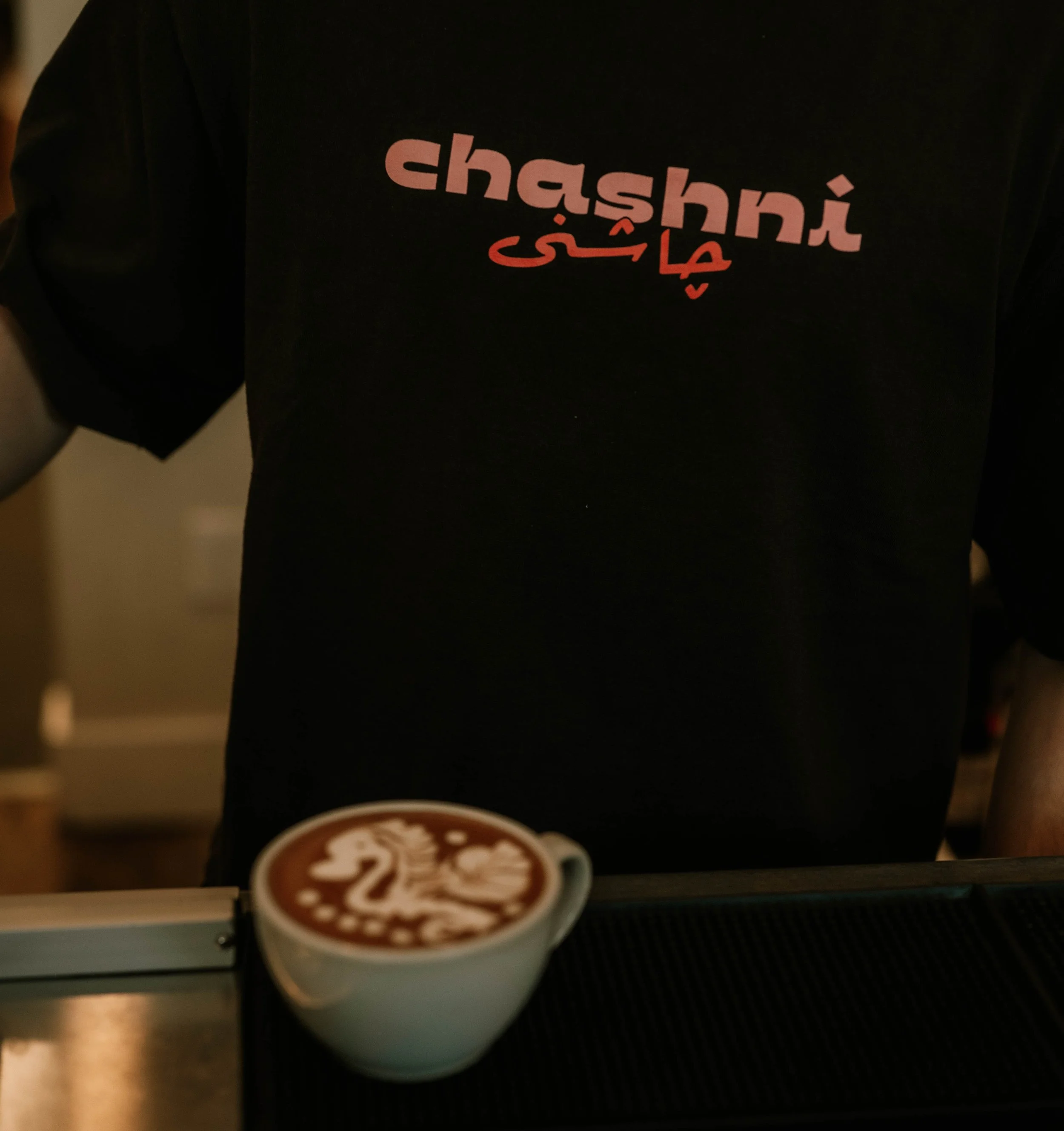 A small coffee cup with a logo featuring a stylized coffee bean and the text '$2' on top of a black surface in a cafe or coffee shop environment.